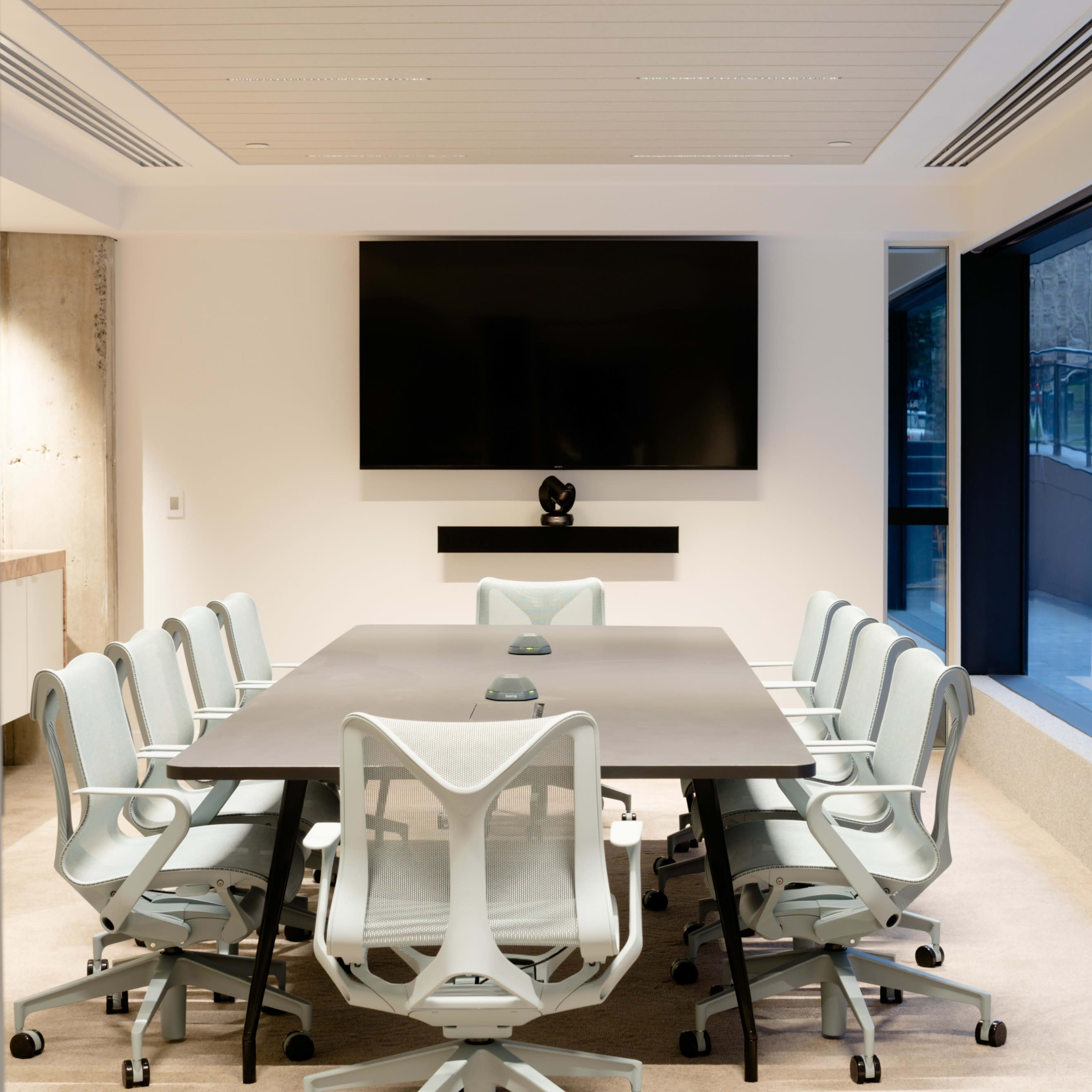 A modern conference room features a long table with twelve ergonomic chairs, a wall-mounted television, and large windows.