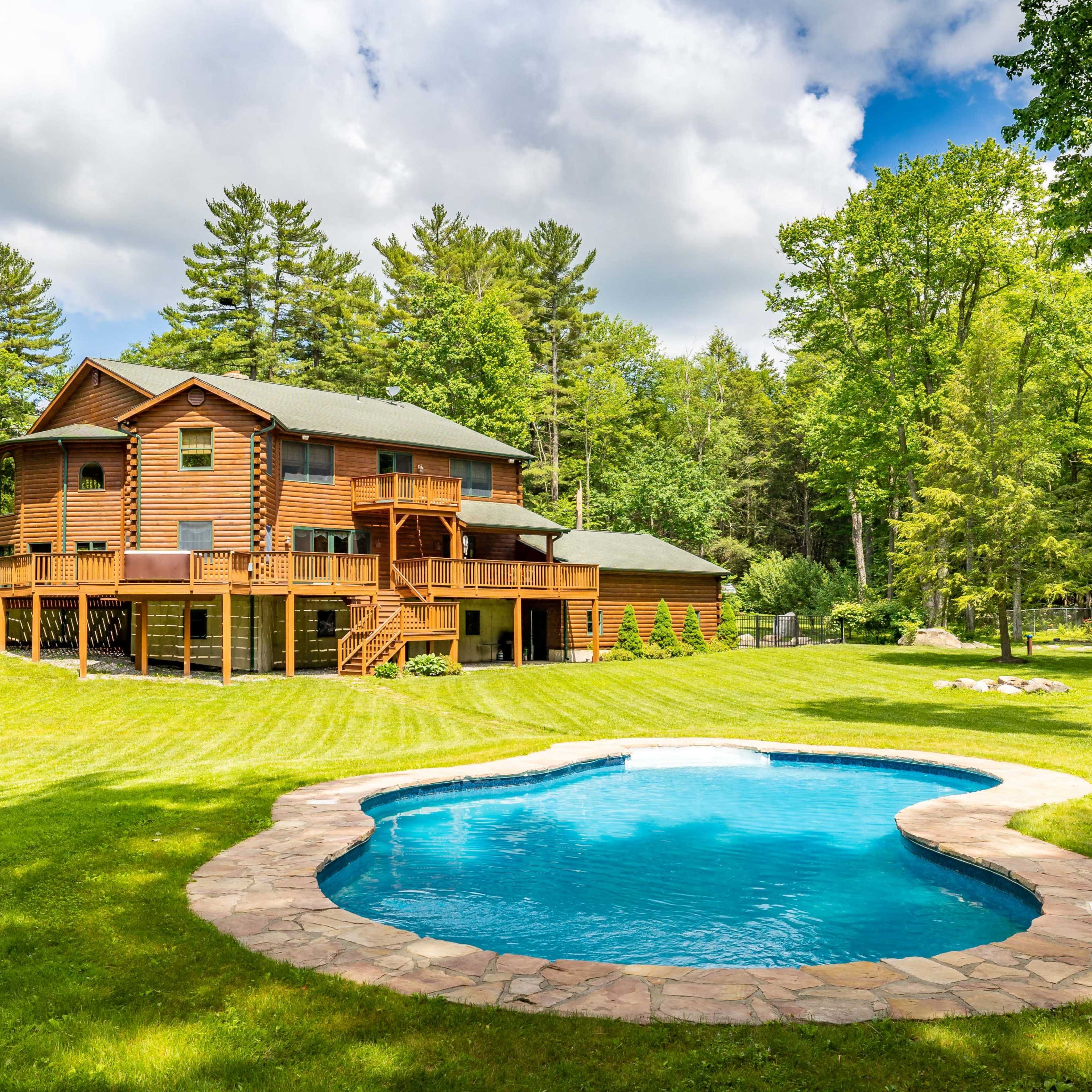 A log cabin-style house with multiple levels and a pool is surrounded by a lush green lawn and trees.