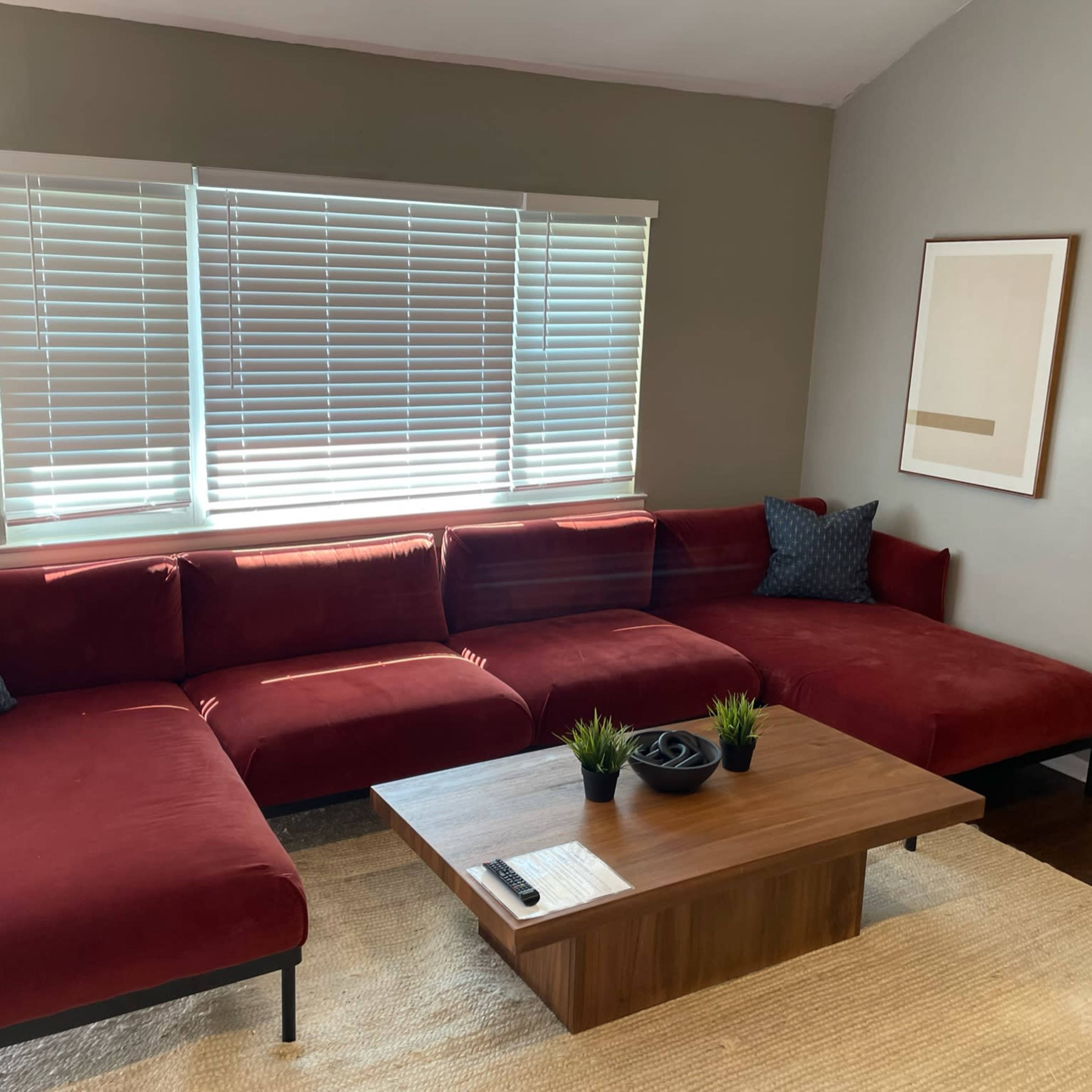 A red sectional sofa with blue accent pillows accompanies a wooden coffee table in a room featuring large windows covered with white blinds.