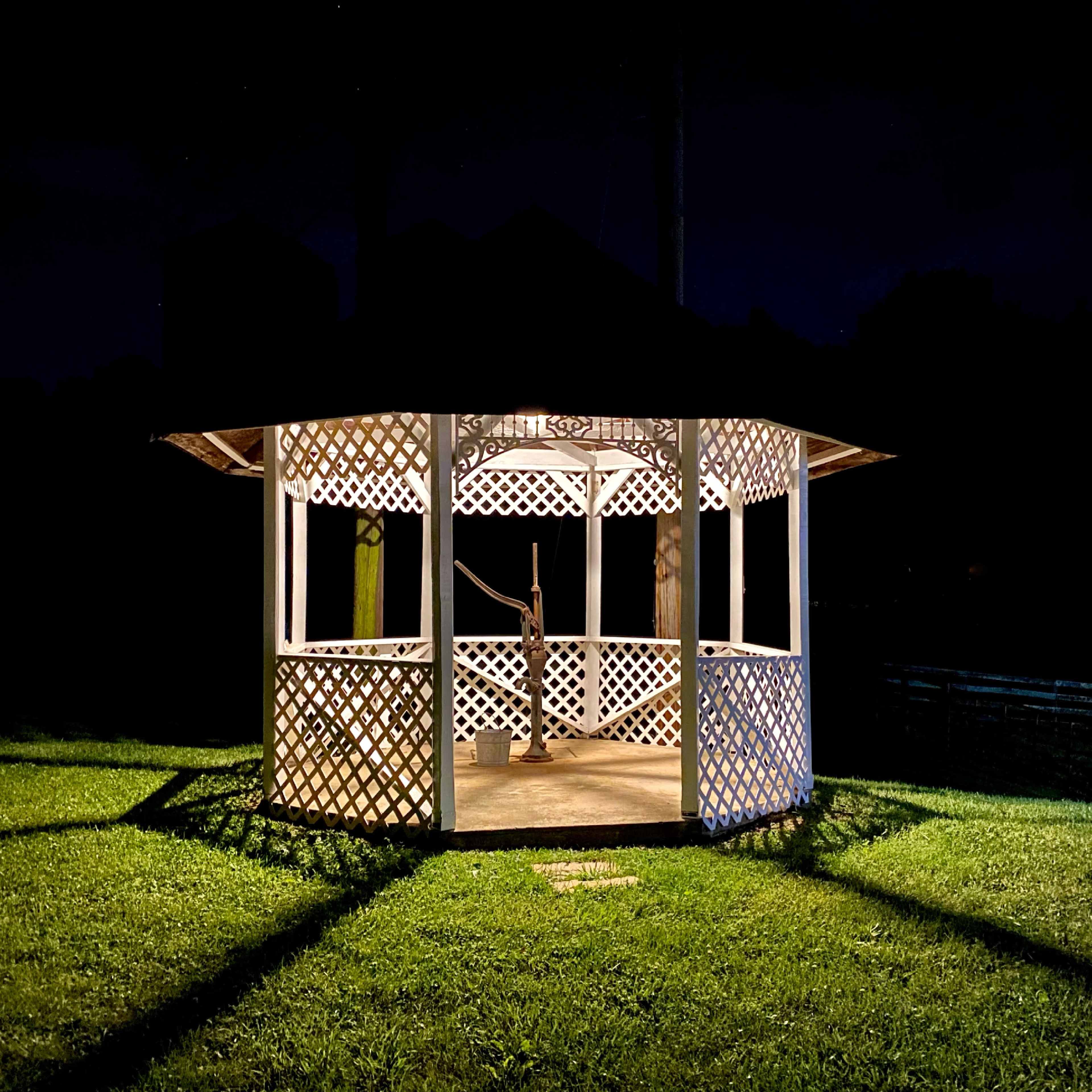 A lighted gazebo stands on a grassy area at night, casting long shadows on the ground.
