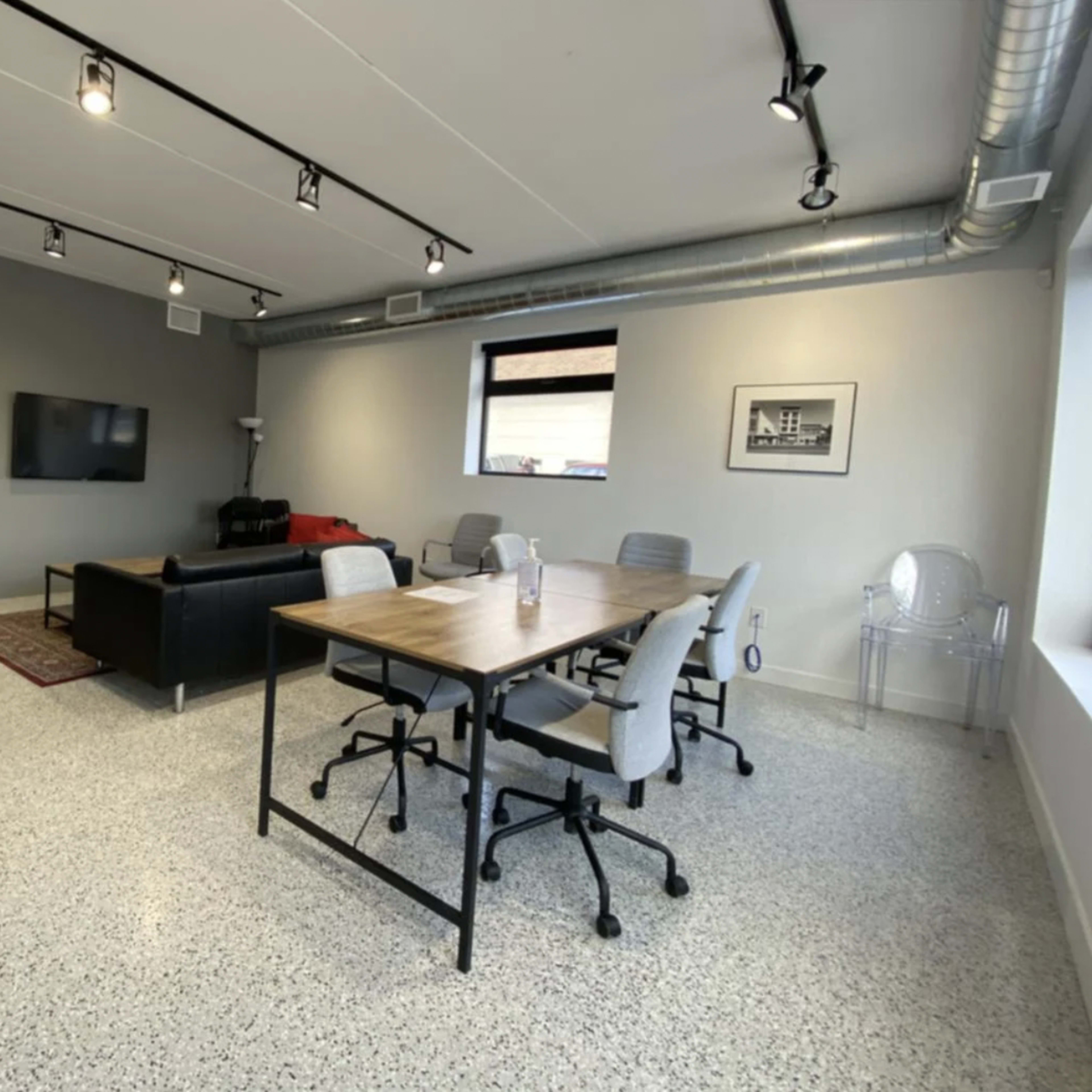 A modern office space featuring a seating area with a black sofa and a wall-mounted TV, along with a conference table surrounded by chairs.