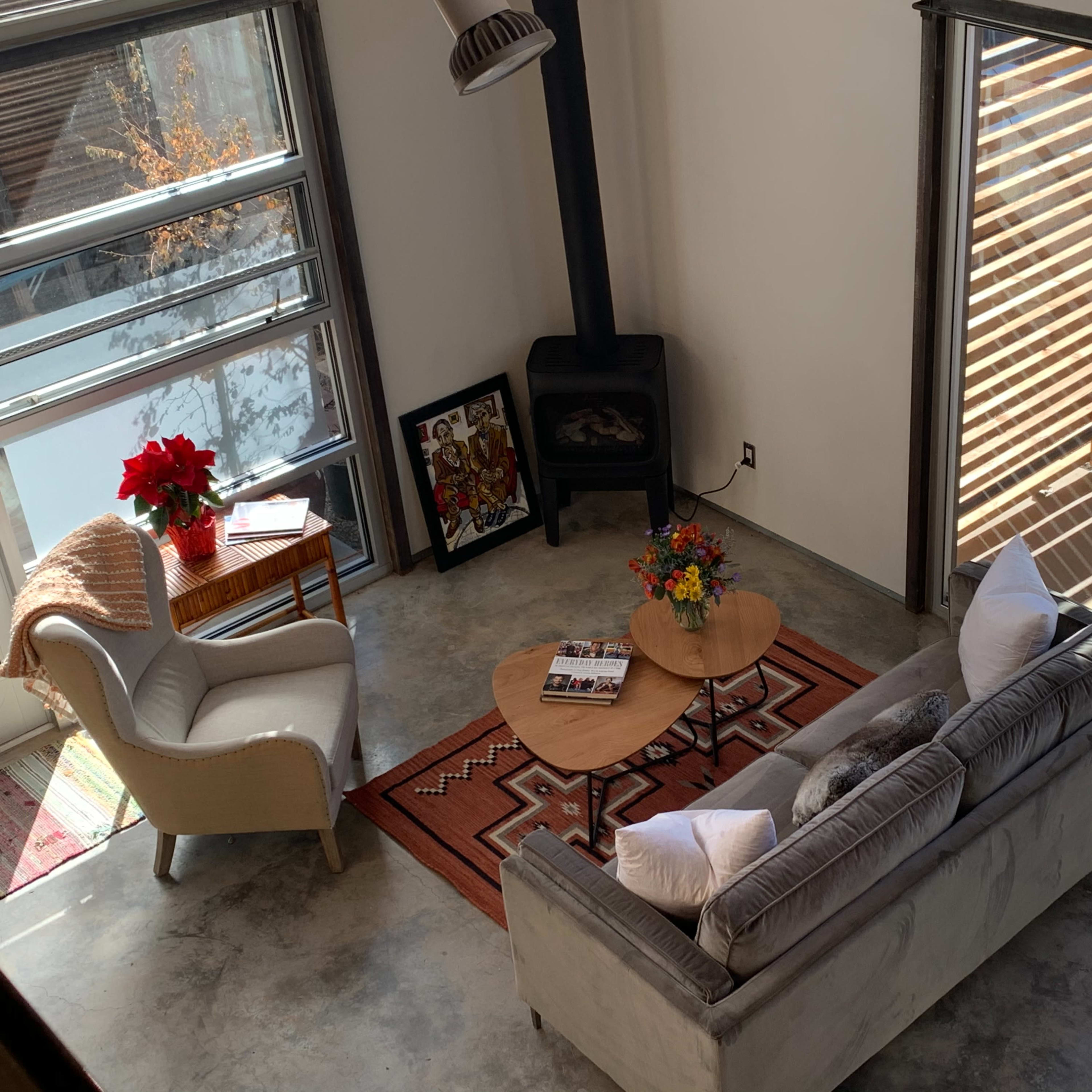 A living room features a gray sofa, a round wooden coffee table, a small side table with a plant, and large glass doors that open to an outdoor space with shade slats.