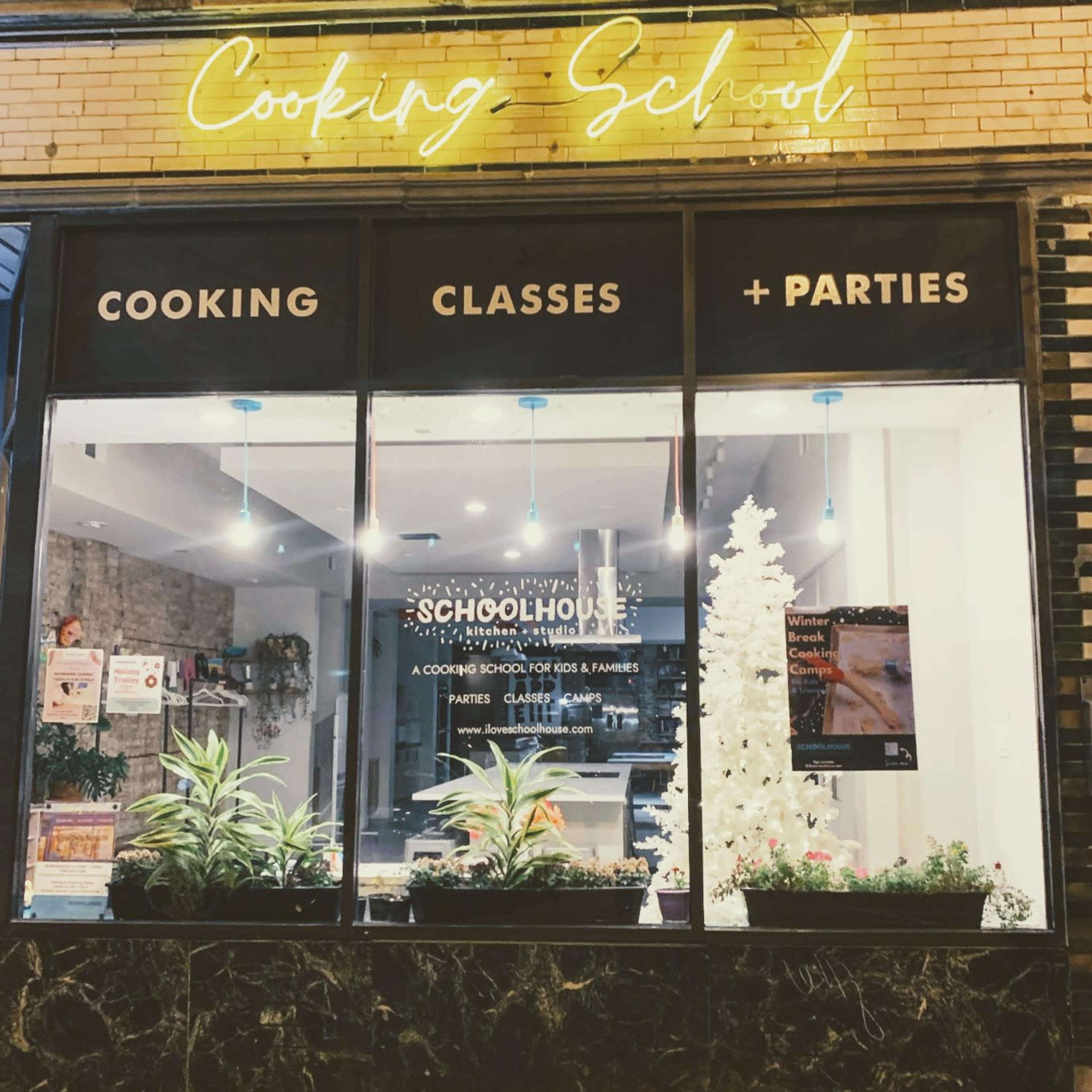 A storefront with a sign that reads "Cooking School" and displays advertisements for cooking classes and parties, along with decorative plants and a white winter-themed display inside.
