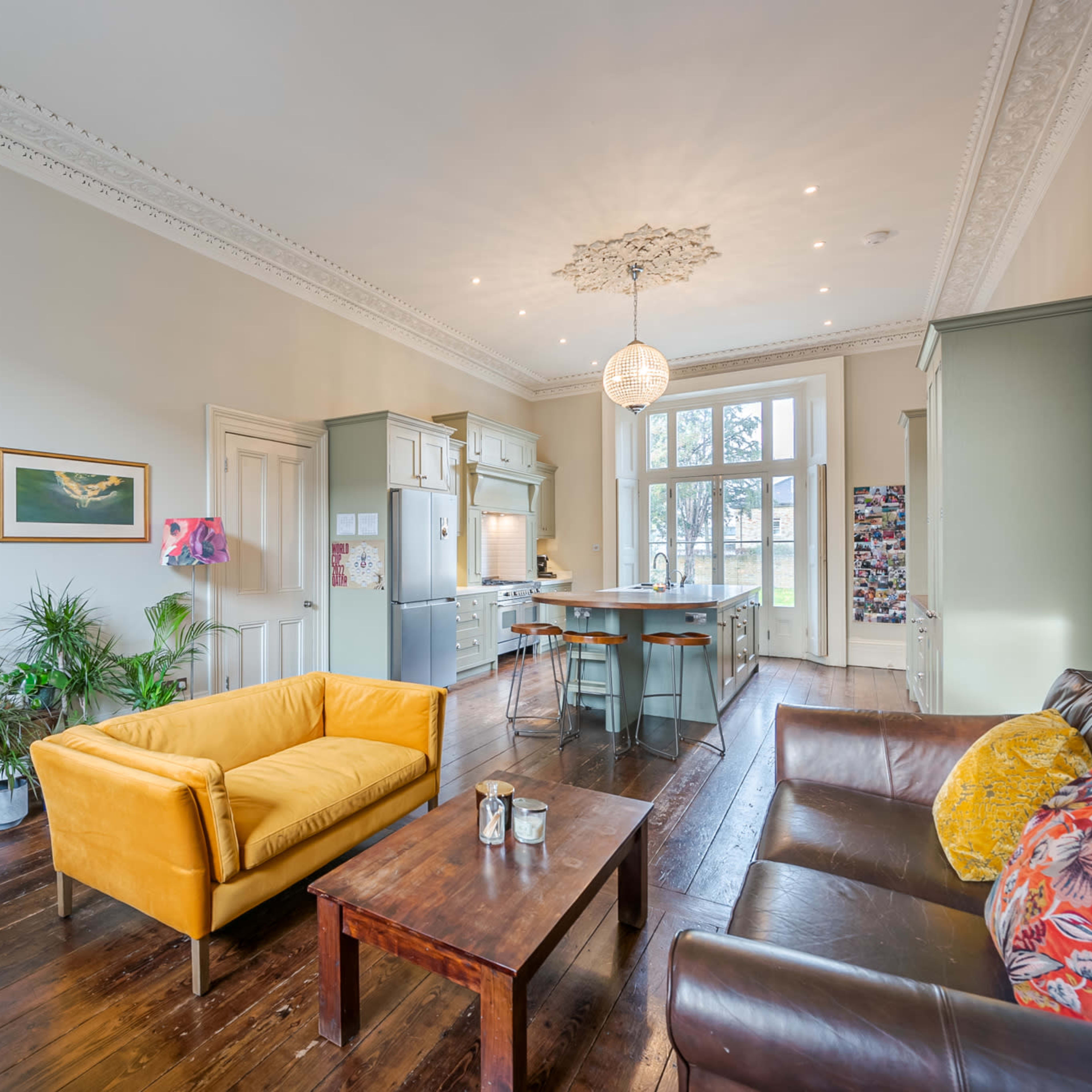A stylish living room with a yellow sofa, a brown leather couch, and a wooden coffee table, leading to a kitchen area with large windows and modern appliances.