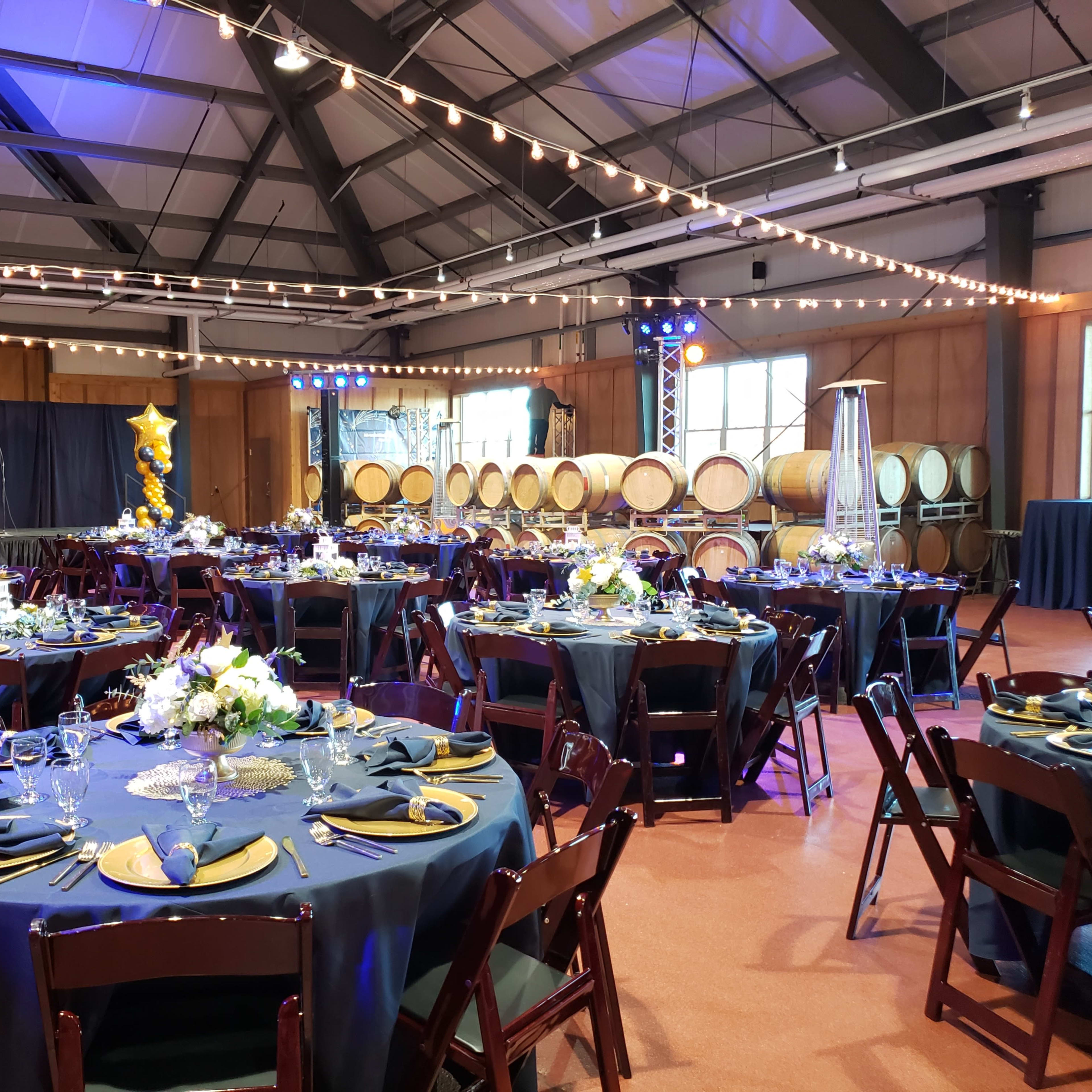A large event space with round tables set for dining, decorated with tableware and centerpieces, alongside wooden wine barrels in the background.