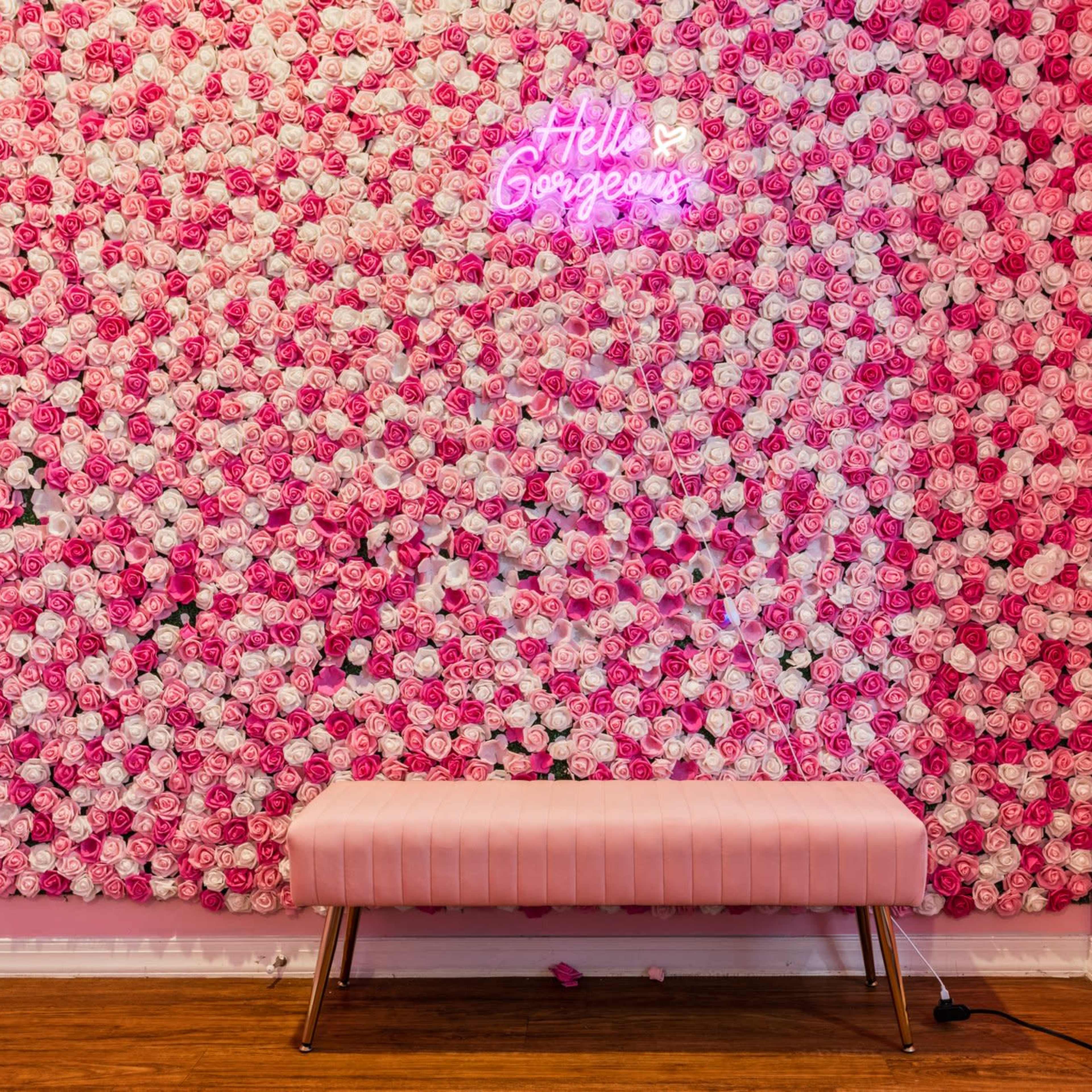 A wall covered in faux roses in various shades of pink, with a neon sign that reads "Hello Gorgeous" and a pink bench in front of it.