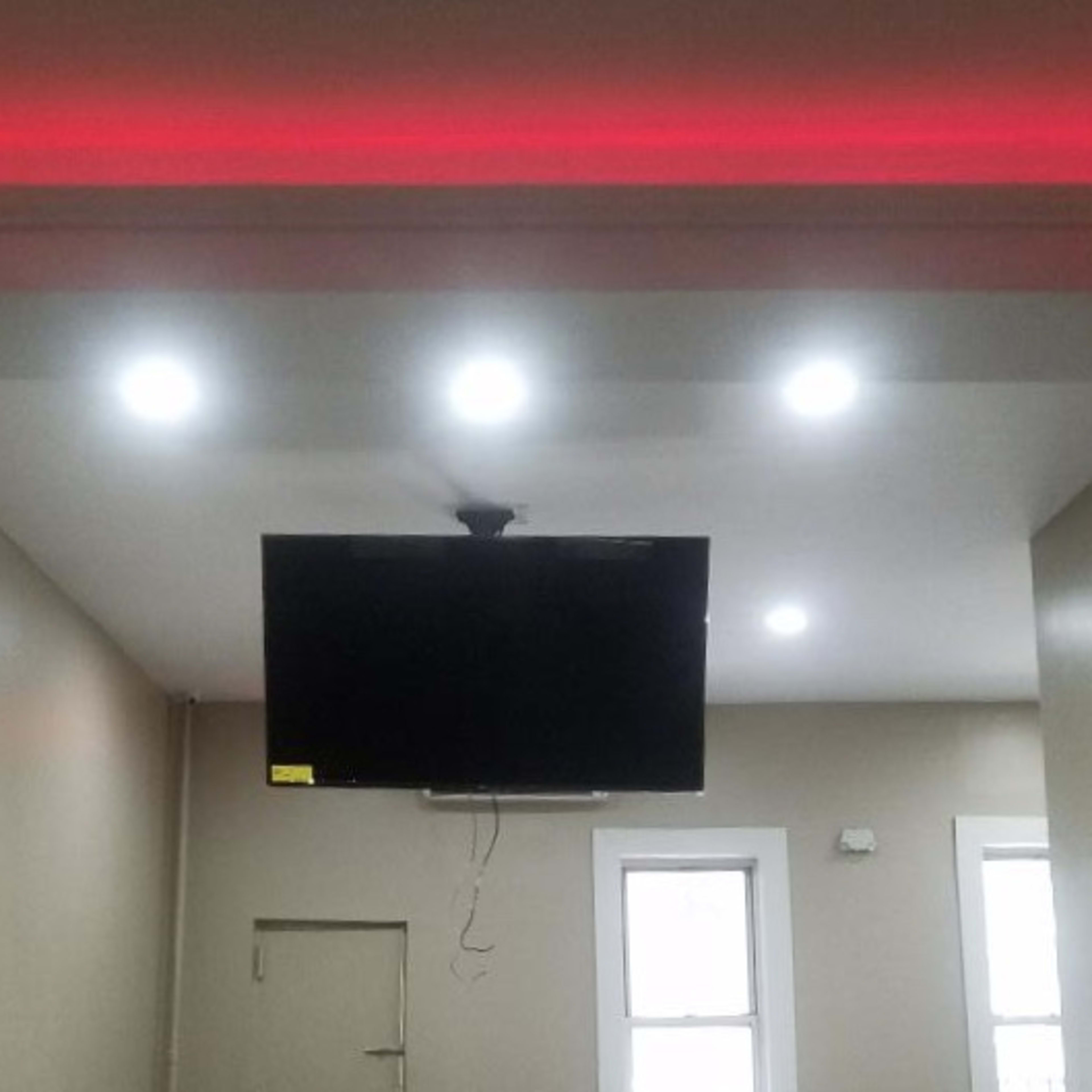 A room with a television mounted on the wall, flanked by recessed lighting and a red-lit ceiling trim.