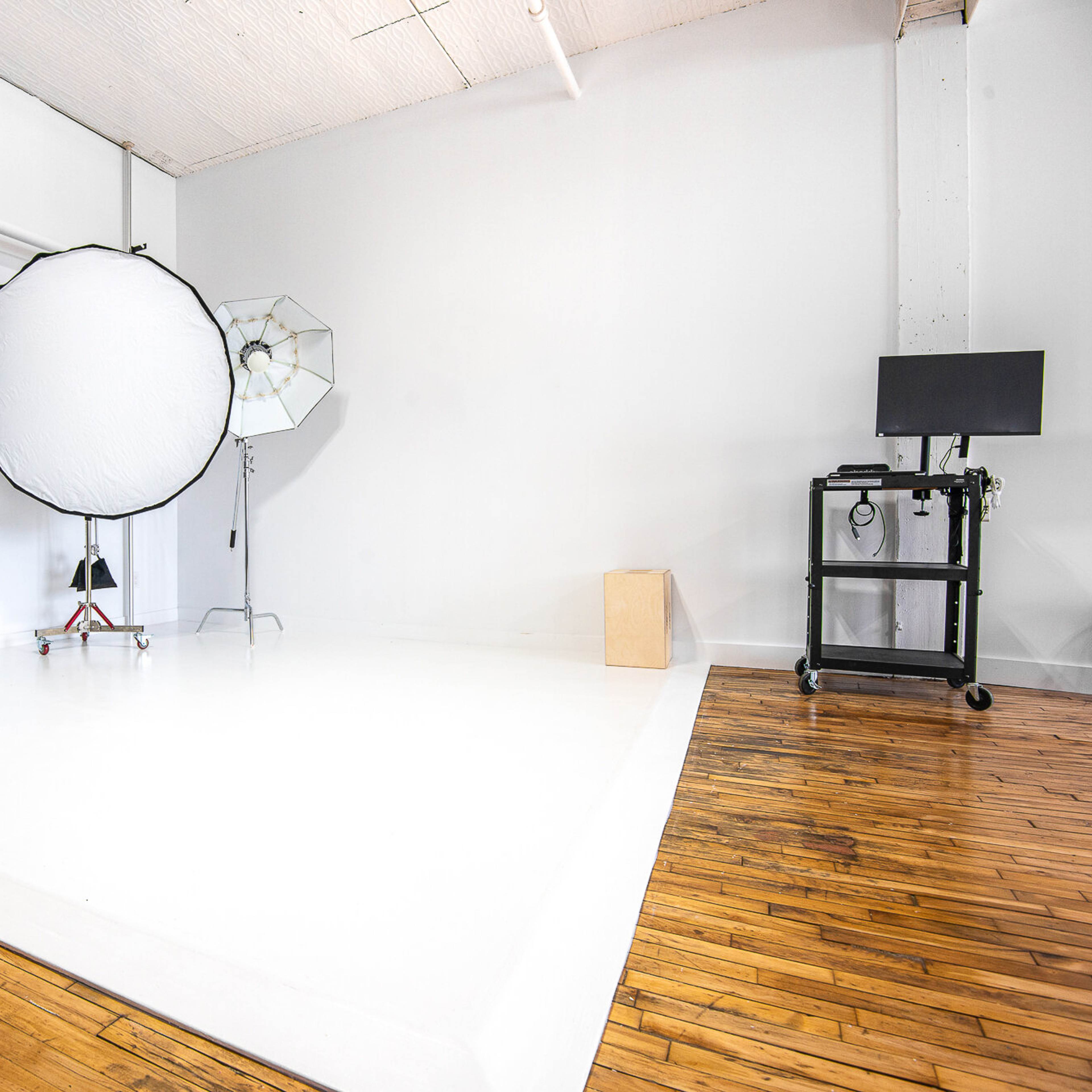 A photography studio with white walls, a polished wooden floor, and various lighting equipment set up, including softboxes and backdrops.