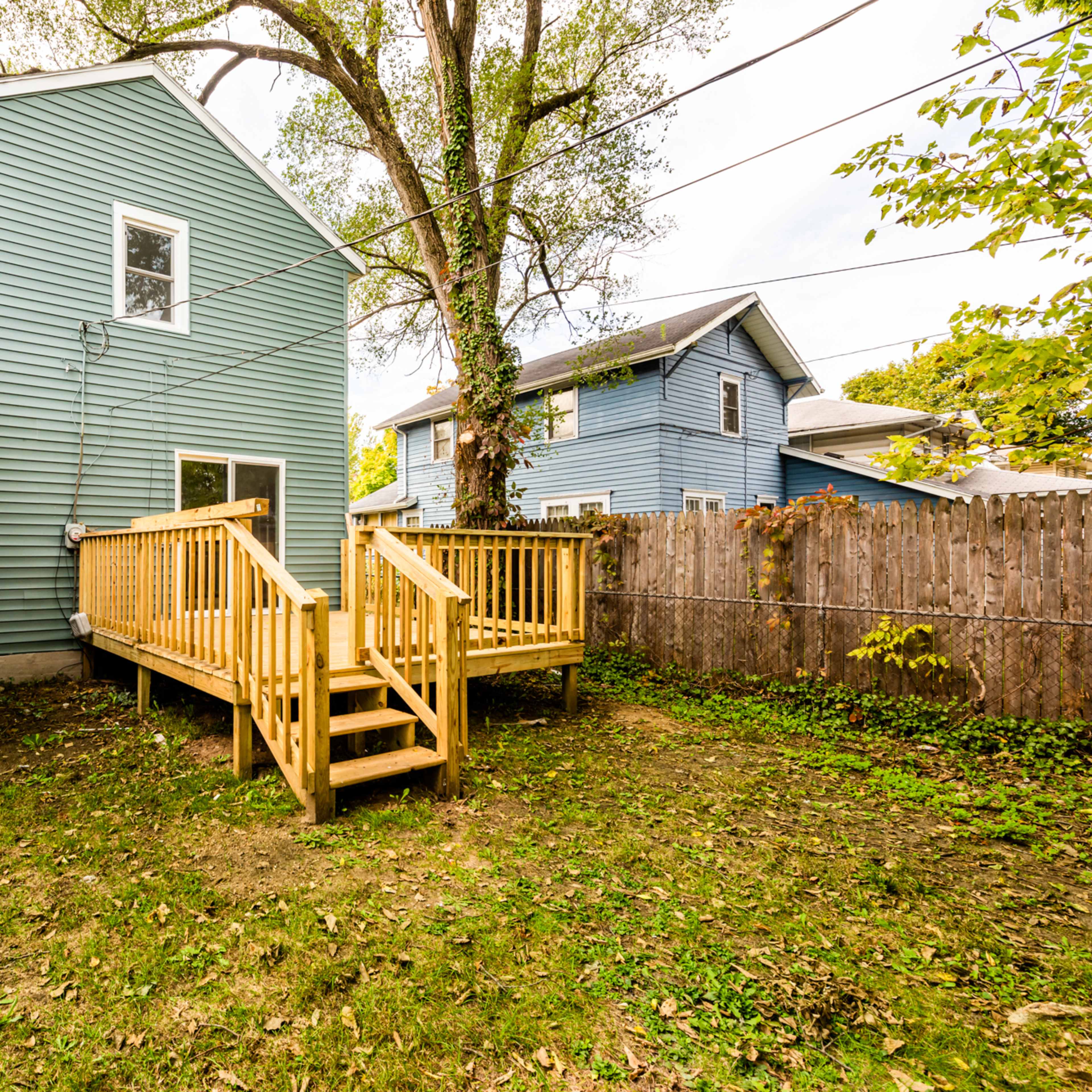 A wooden deck with steps leads to the back entrance of a blue house, surrounded by grass and bordered by a wooden fence.