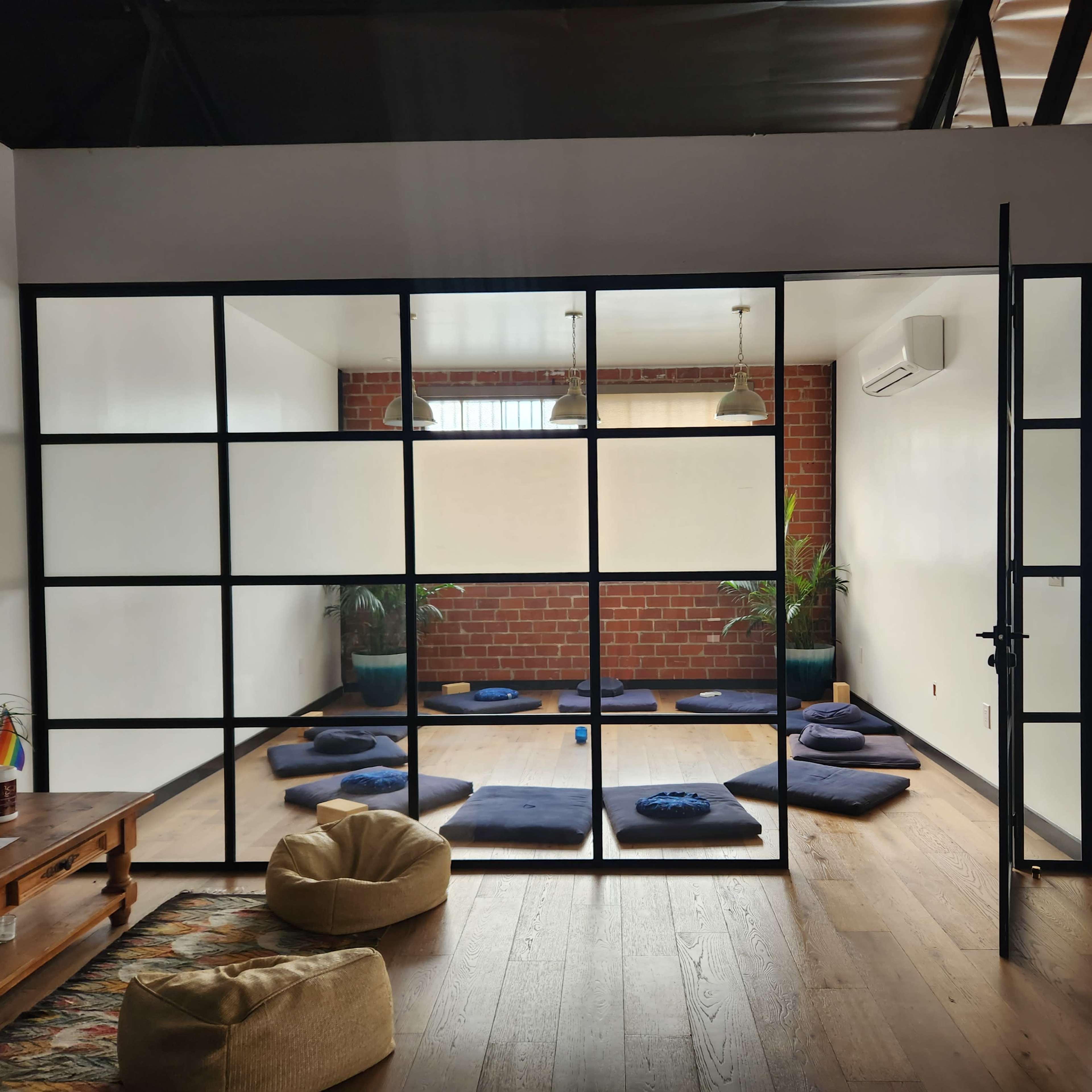 A spacious room divided by a glass panel, featuring blue cushions arranged on the floor and green plants in the corners.