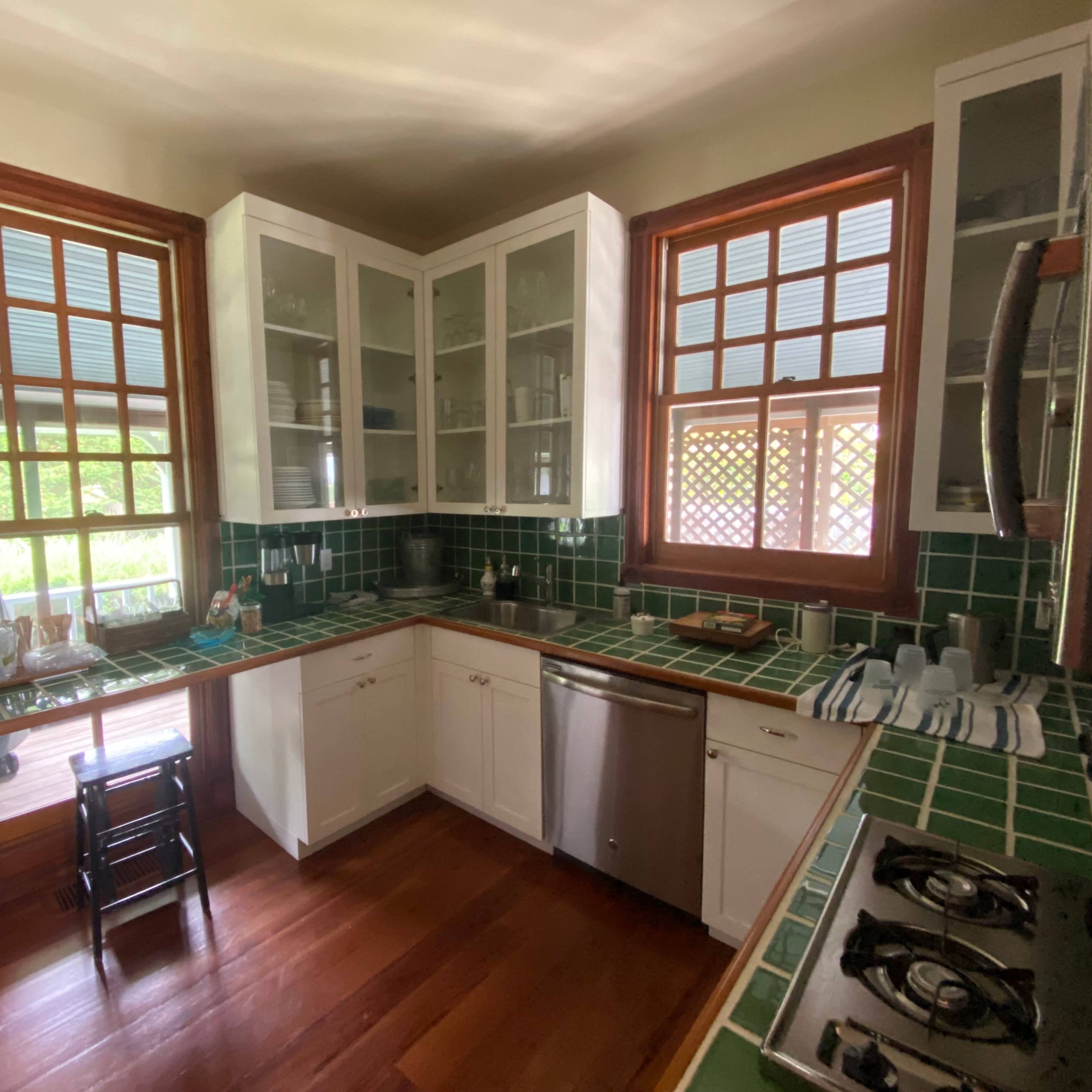 A kitchen with green tiled countertops, white cabinetry, and two large windows letting in natural light.