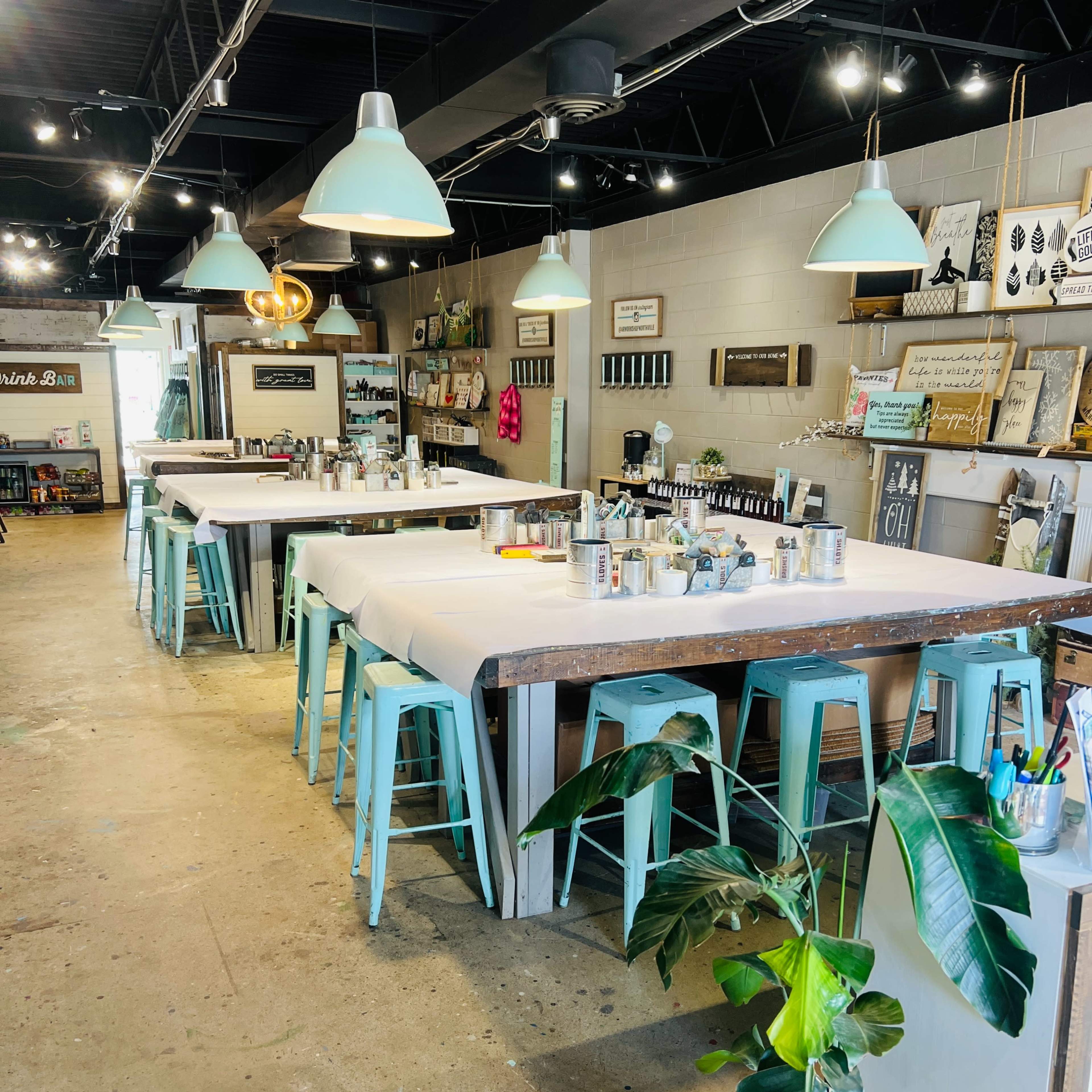 A spacious, well-lit creative studio with several large tables surrounded by turquoise chairs and various art supplies displayed around the room.