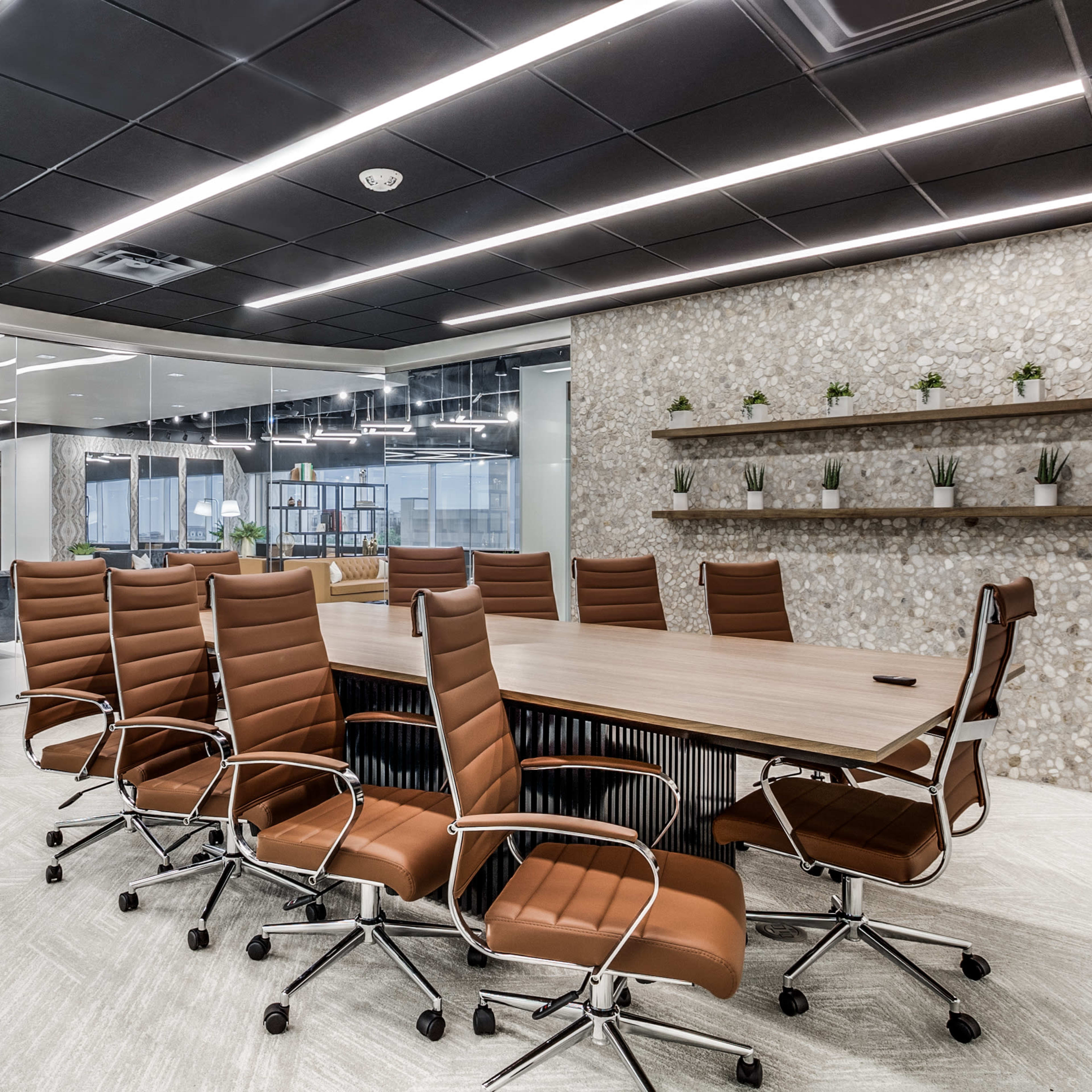 A modern conference room features a long wooden table surrounded by brown leather chairs, with a glass wall and a plant in the corner.