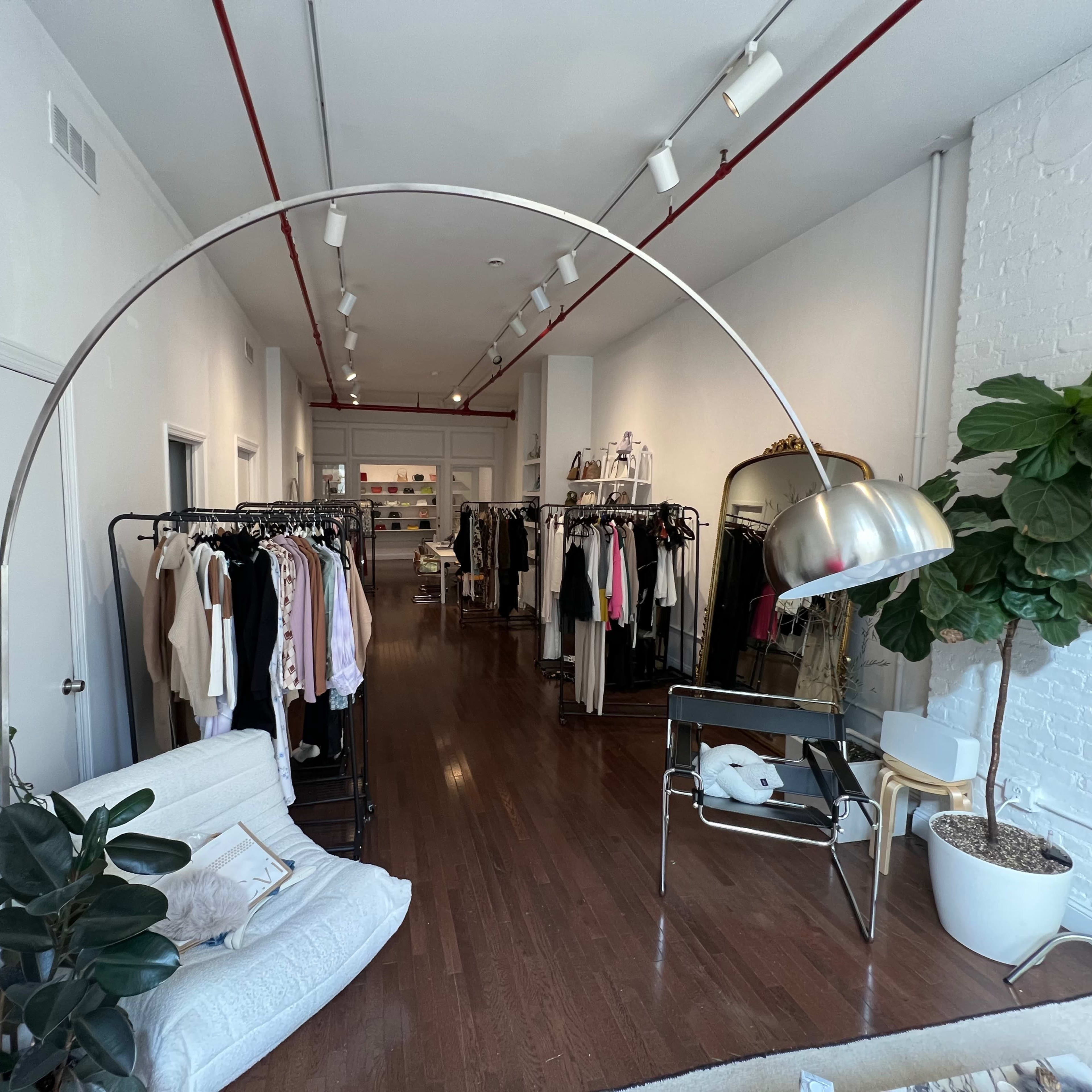 A clothing store interior with racks of clothes on either side, a floor lamp arching over a seating area, and large windows allowing natural light to enter.