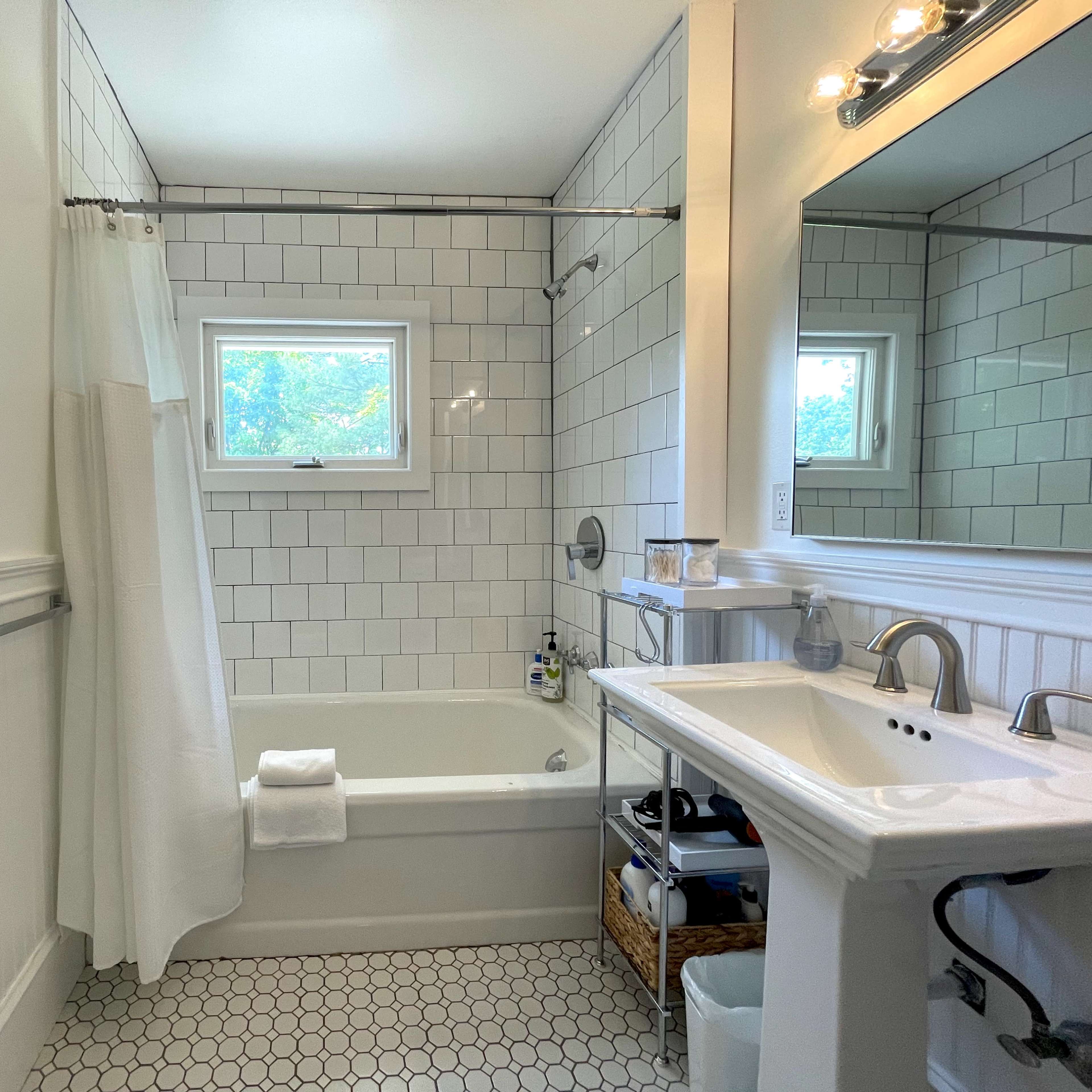 A bathroom featuring a white tiled wall, a bathtub with a shower, a sink, and a window providing natural light.