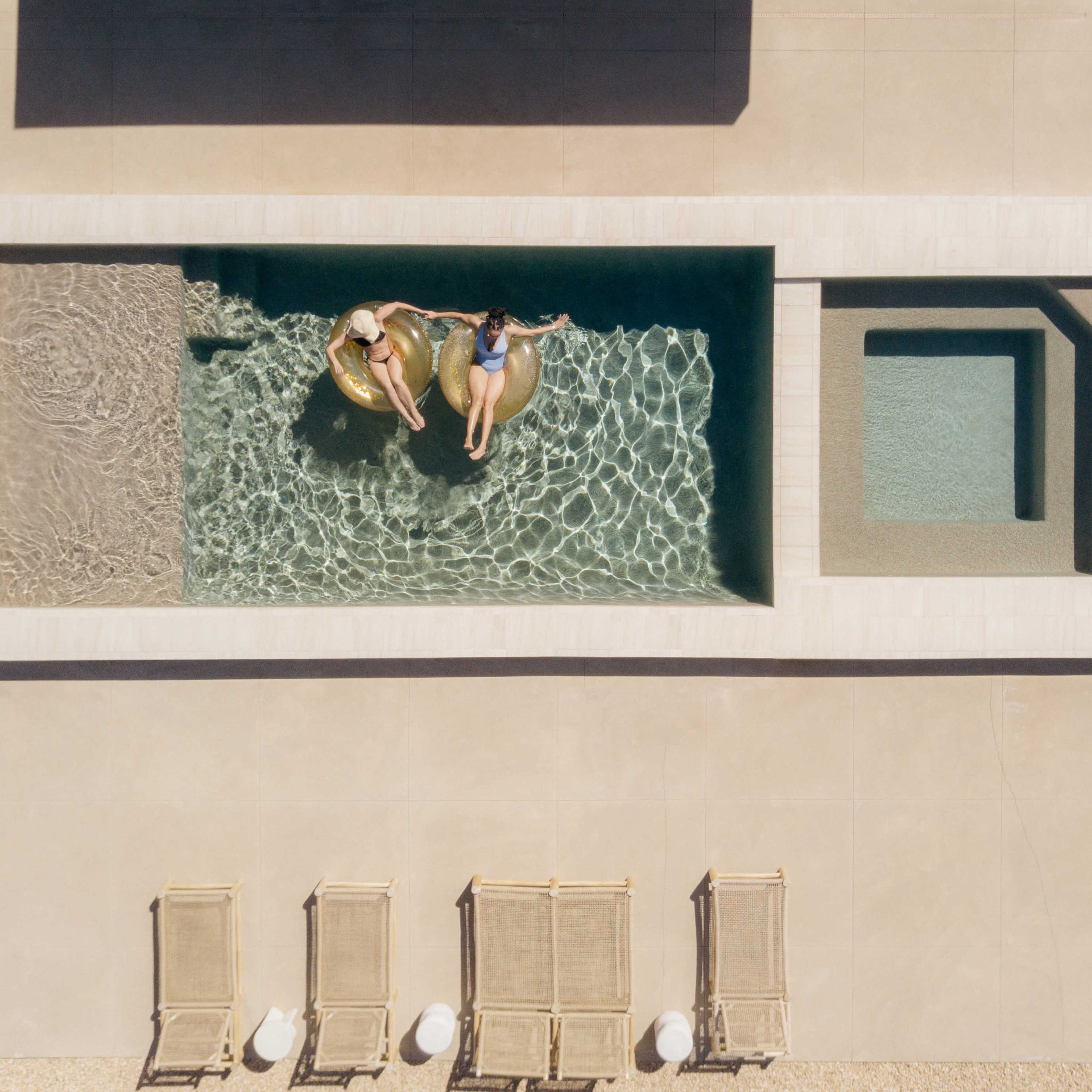 Two individuals relax on floatation devices in a rectangular swimming pool, surrounded by lounge chairs and a spa area.