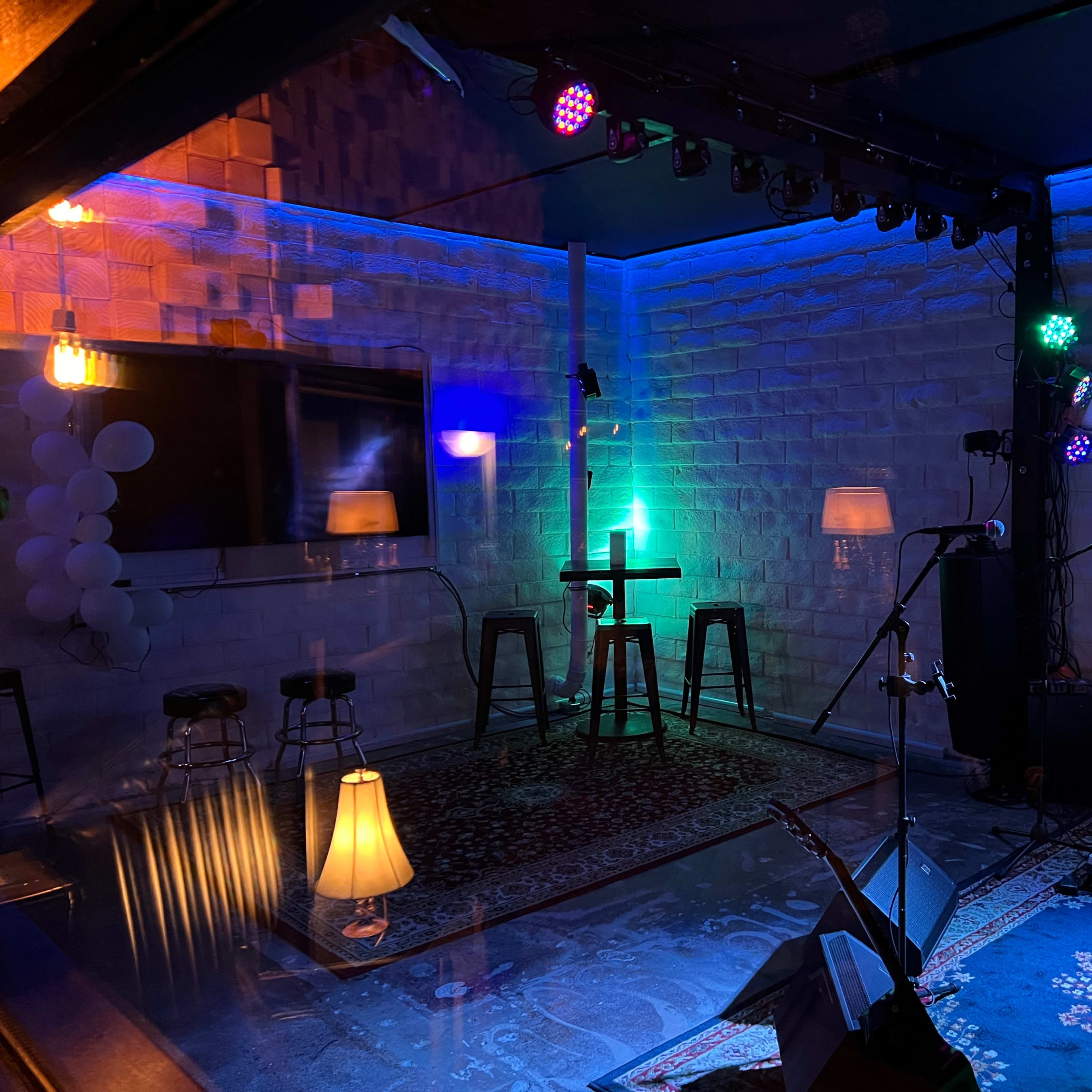 A small performance space with colorful lighting, several stools, and musical equipment, featuring a patterned rug and floor lamps.
