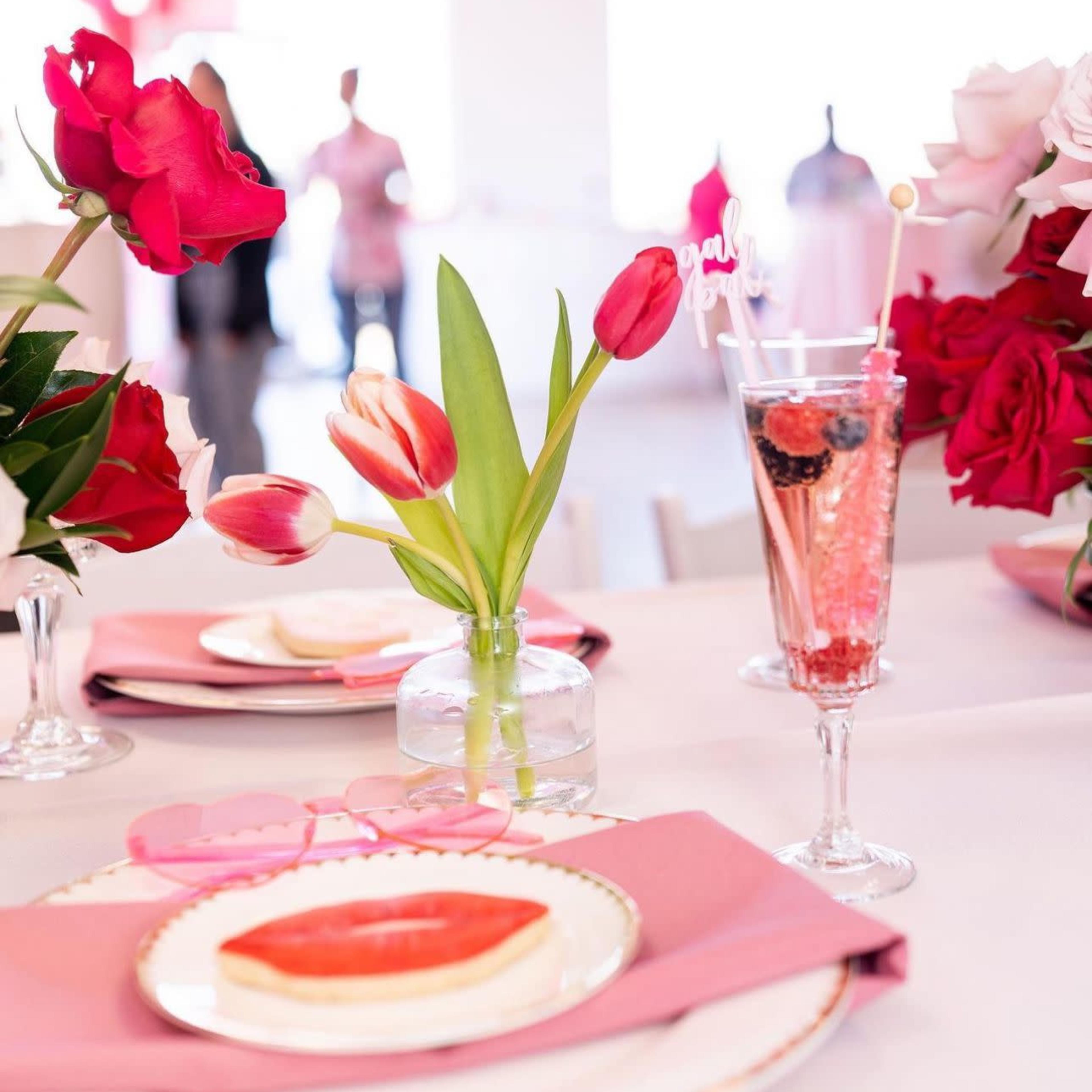 The table is set with pink napkins, a vase of tulips, and a pink drink adorned with fruit, surrounded by red and pink floral arrangements.