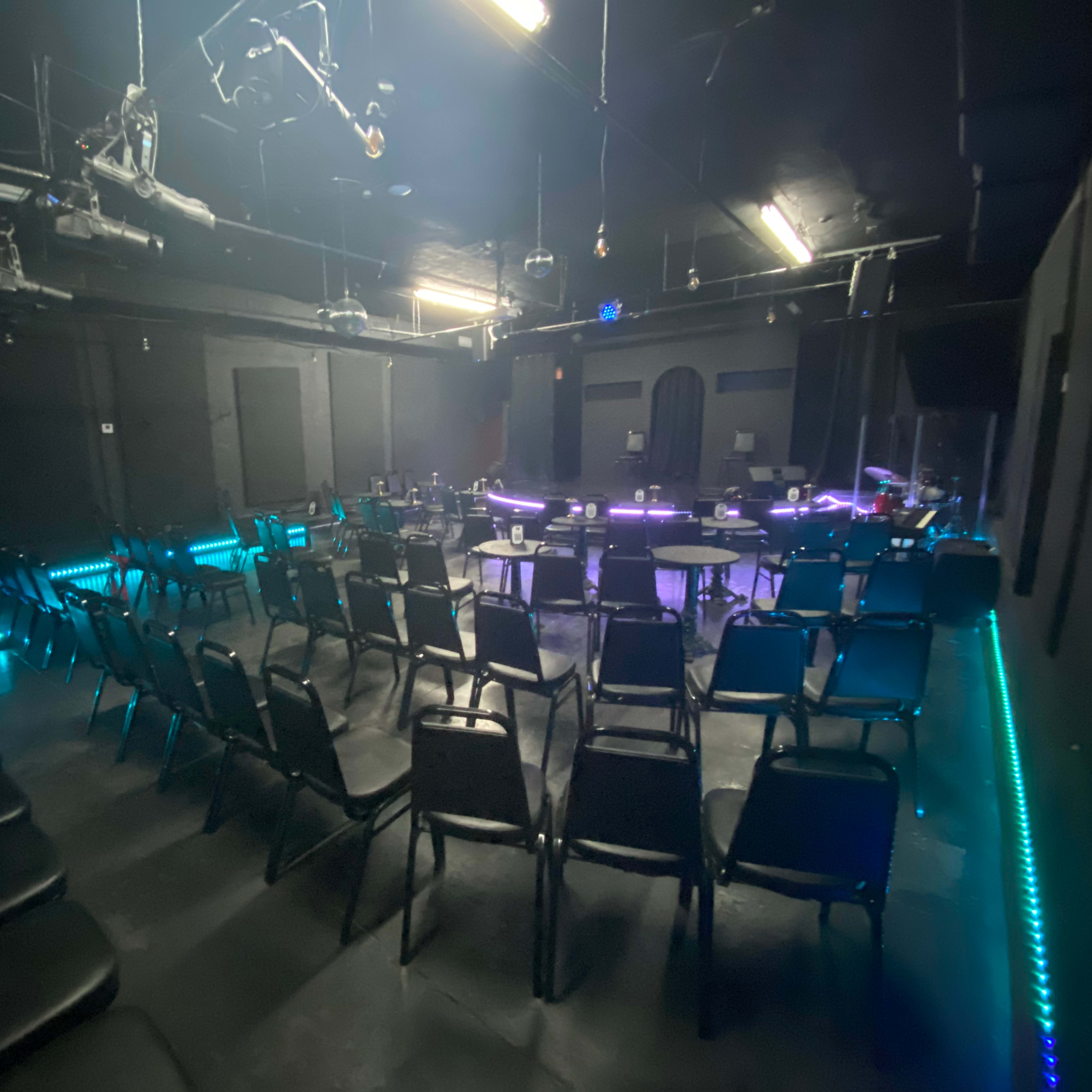 A dimly lit performance space with rows of black chairs arranged in front of a small stage adorned with colorful lights.