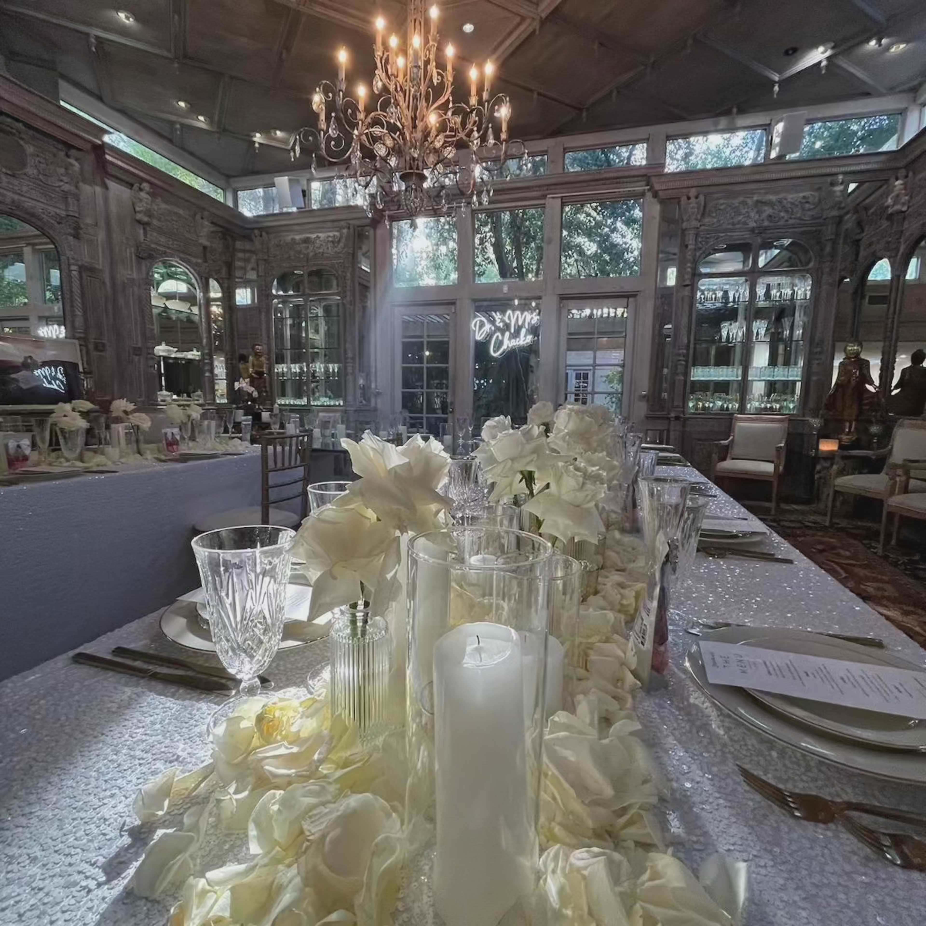 An elegantly set dining table adorned with white flowers and candles, surrounded by a lavishly decorated interior featuring large windows and a chandelier.