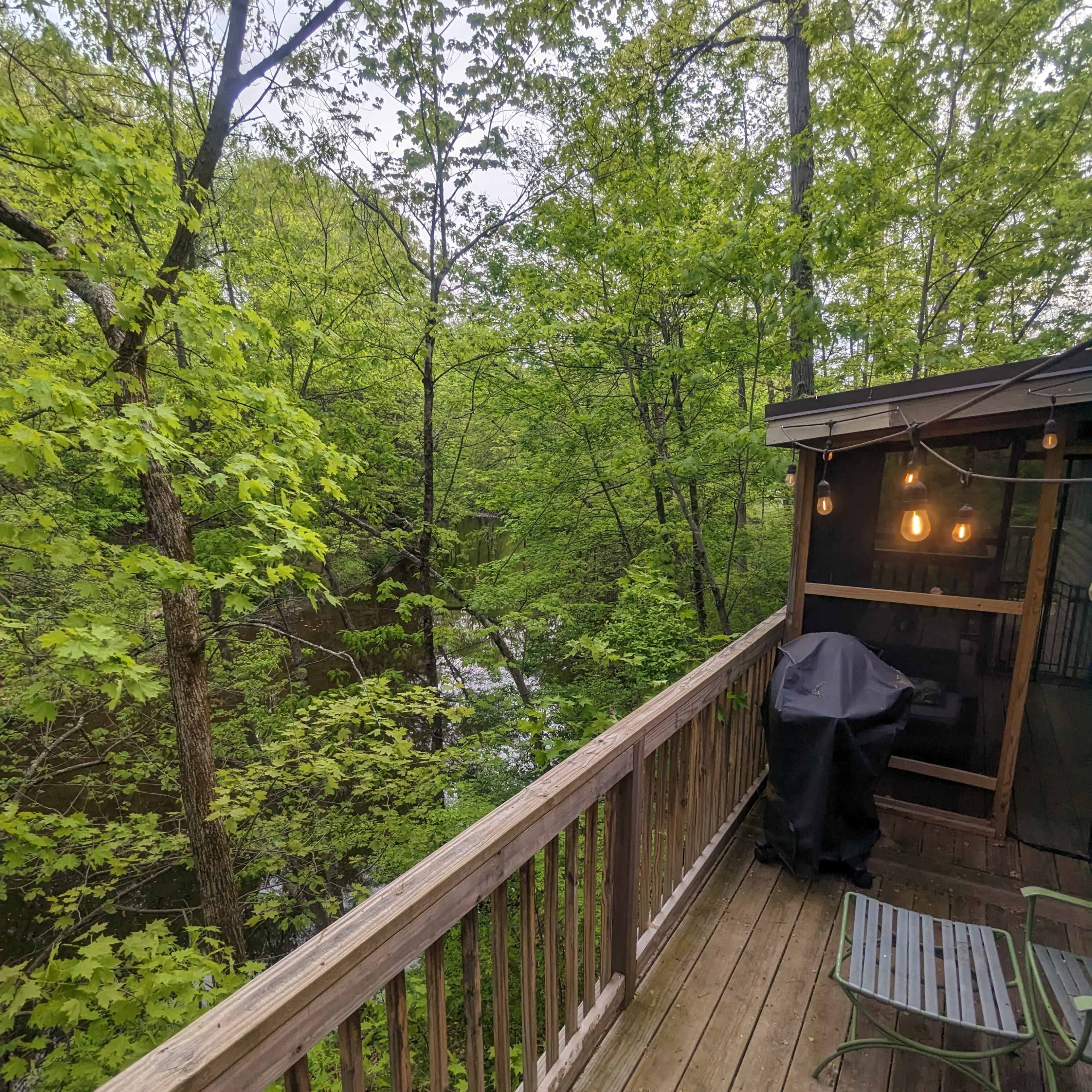 A wooden deck overlooks a leafy green forest and a flowing stream, with a covered grill and a chair positioned beside it.
