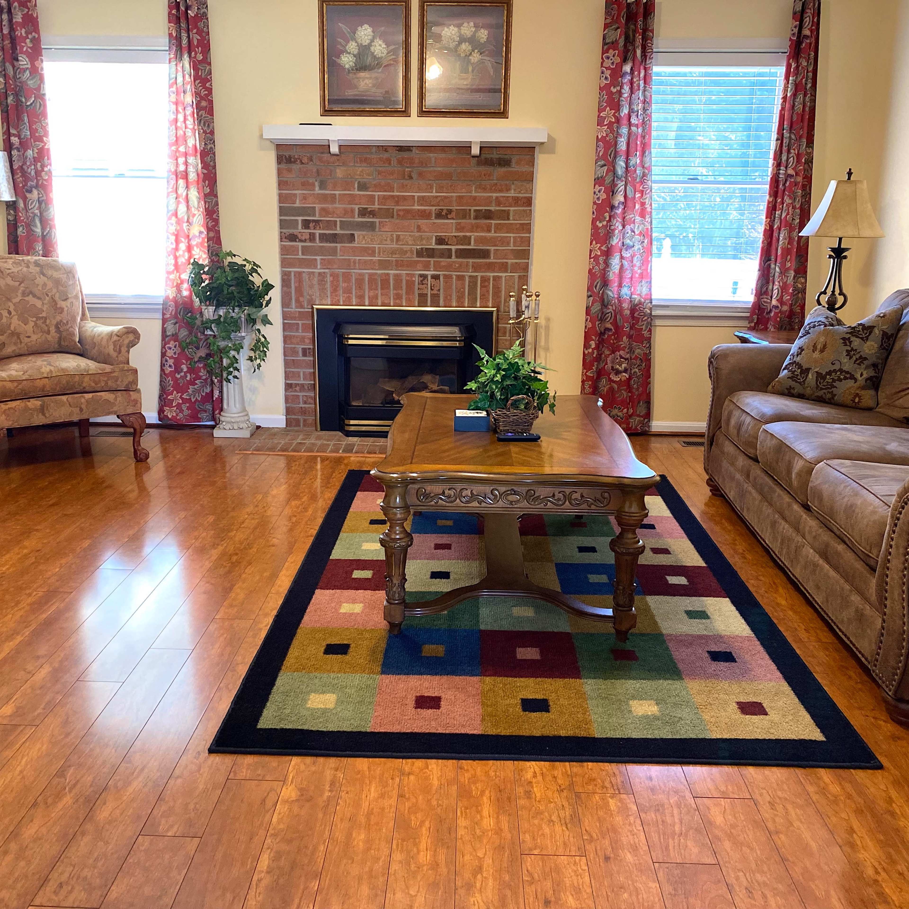 The living room features a brown sofa and armchair, a wooden coffee table, and a fireplace surrounded by decorative plants and framed pictures.