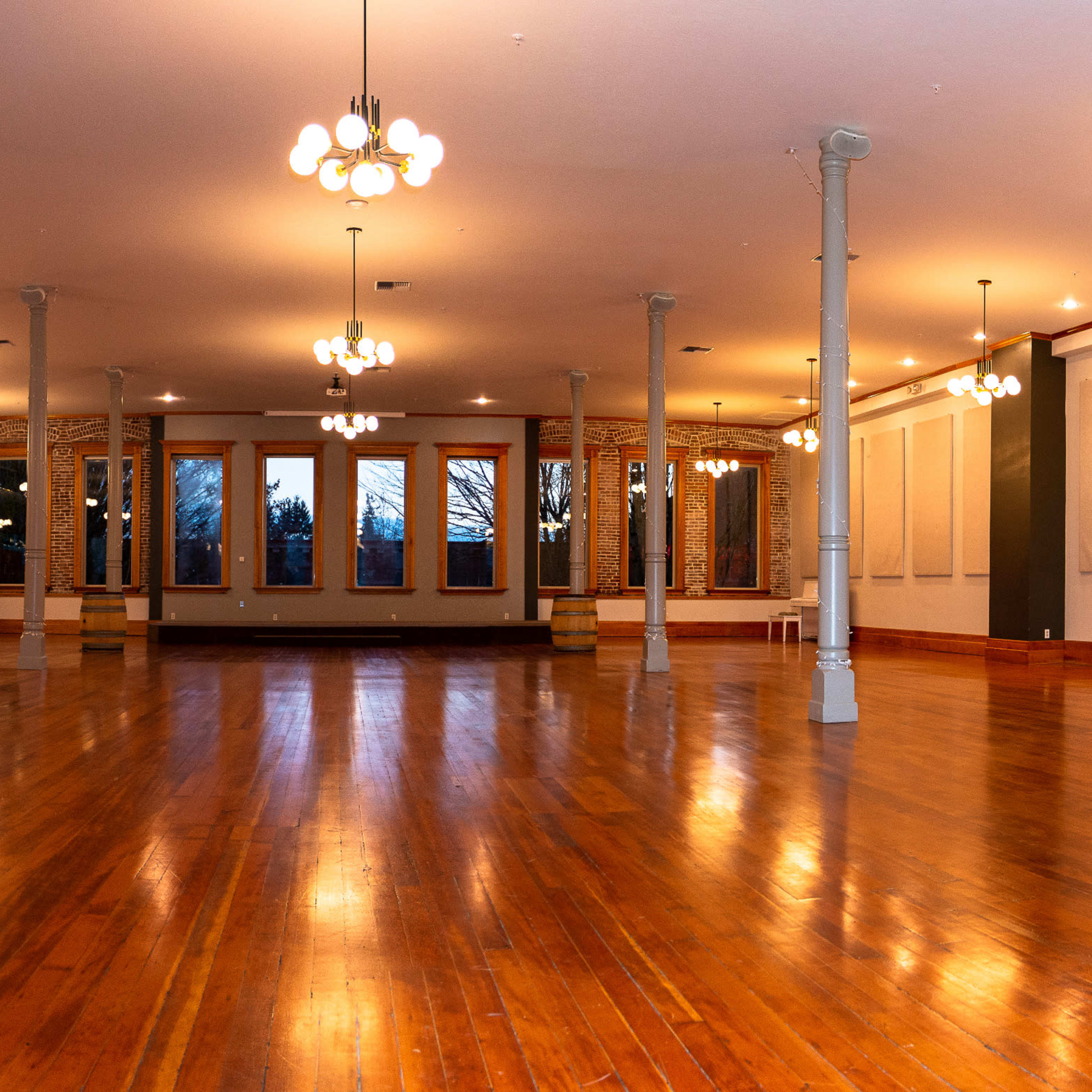 An empty, well-lit event space with wooden flooring, tall ceilings, and large windows.
