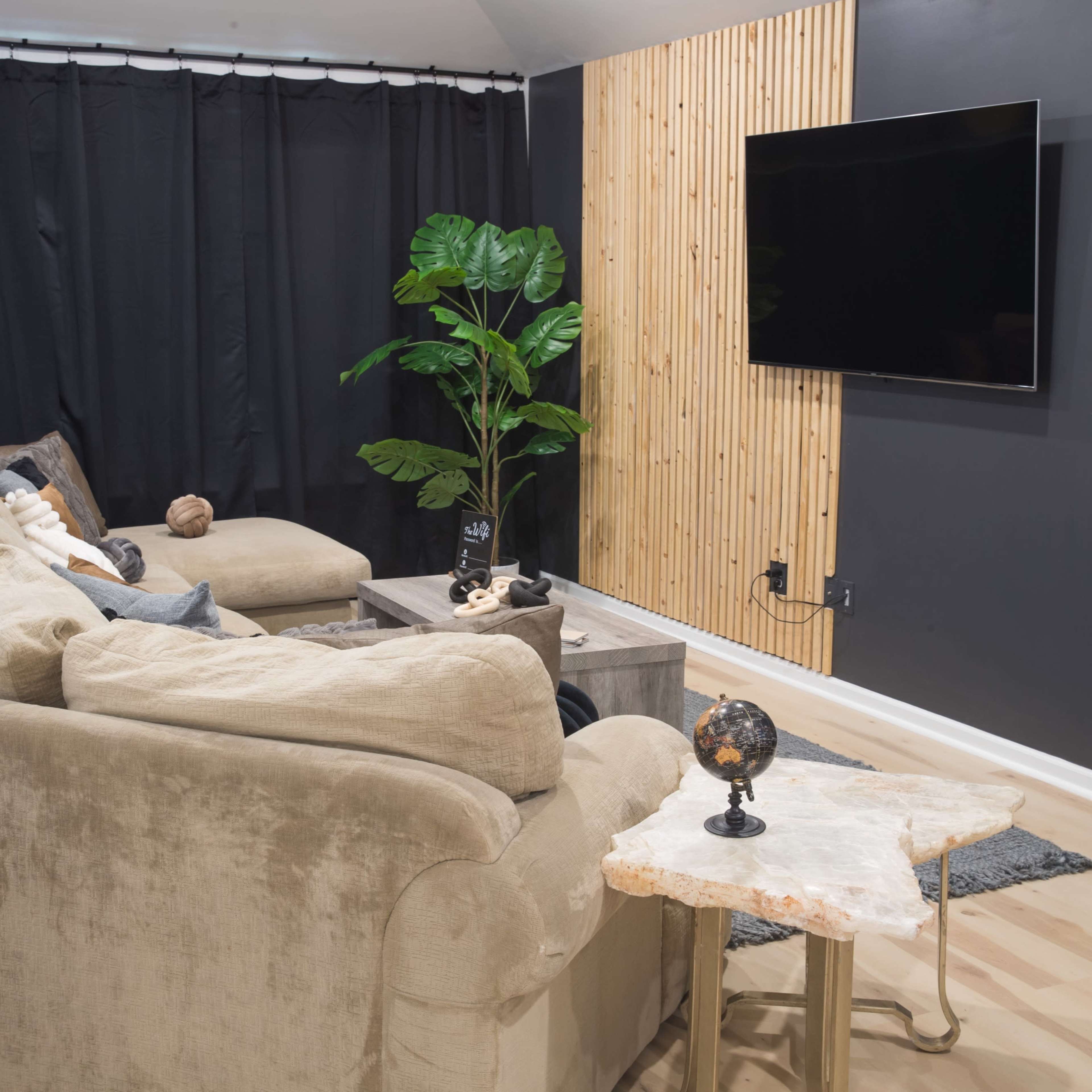 A modern living room with a large, beige sectional sofa facing a wall-mounted television, accompanied by a small side table and a potted plant.
