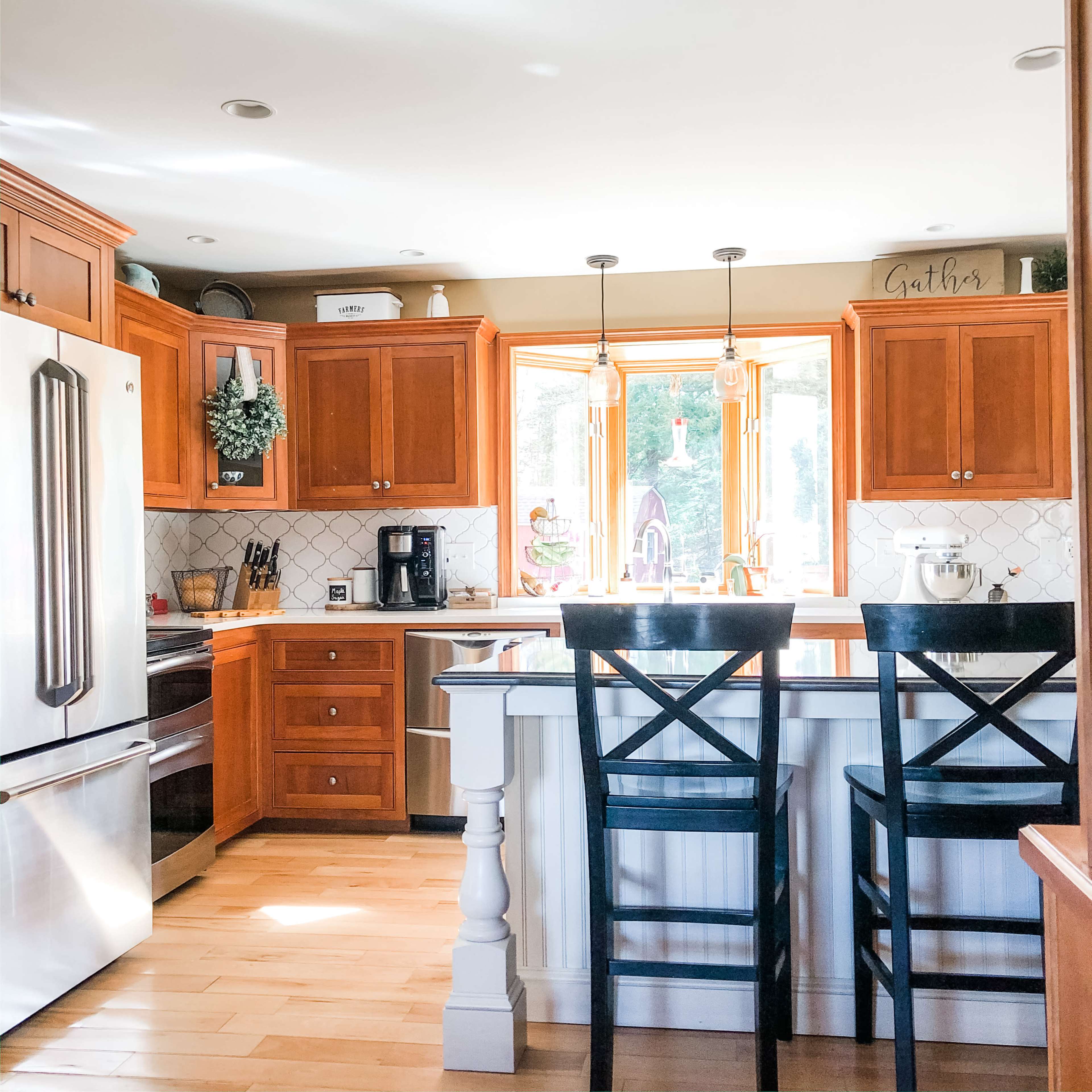 A kitchen with wooden cabinets, a stainless steel refrigerator, and two black bar stools at a white island, with a window displaying natural light.