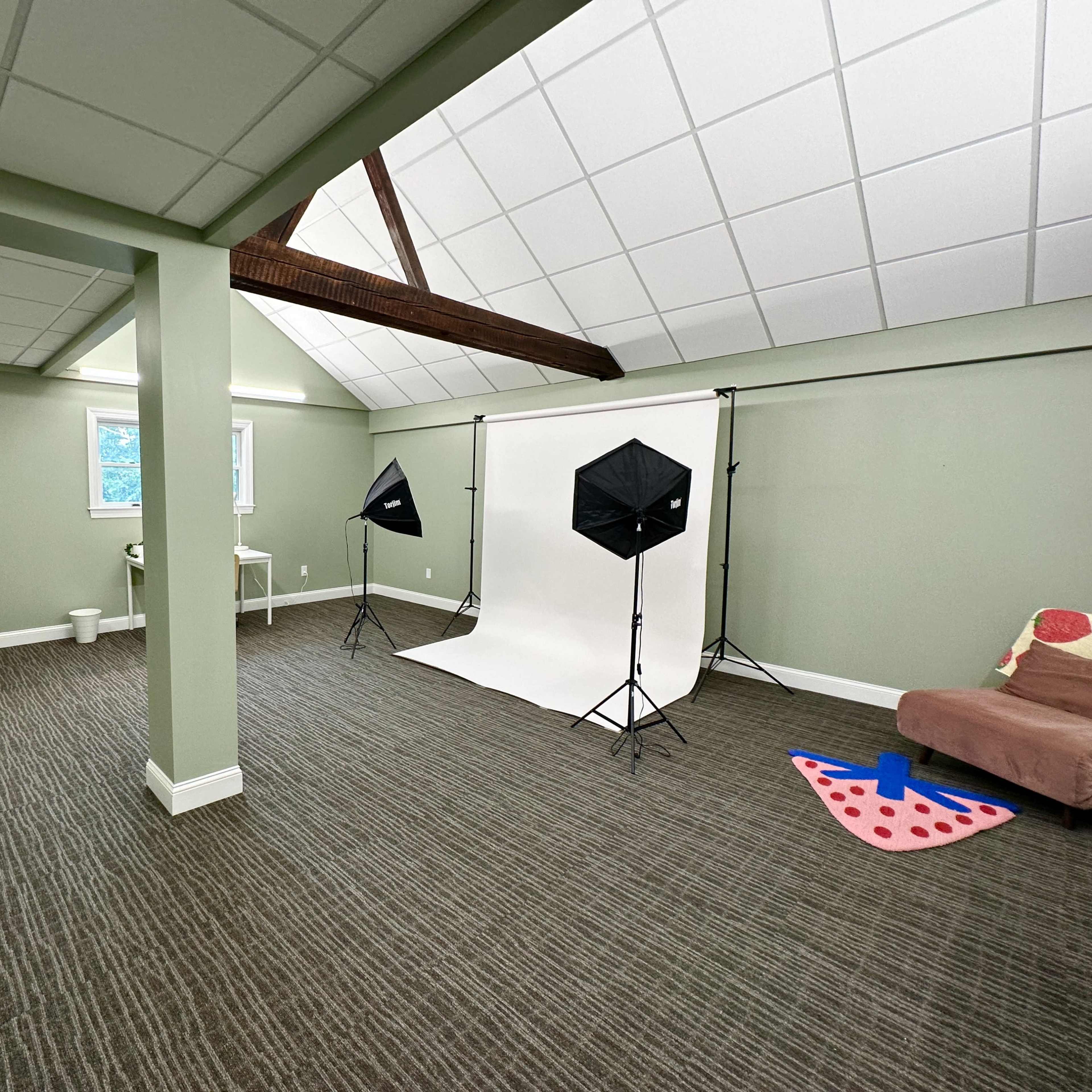 A spacious photo studio with a white backdrop, two softbox lights, a couch, clothing racks, and a colorfully patterned mat on the floor.