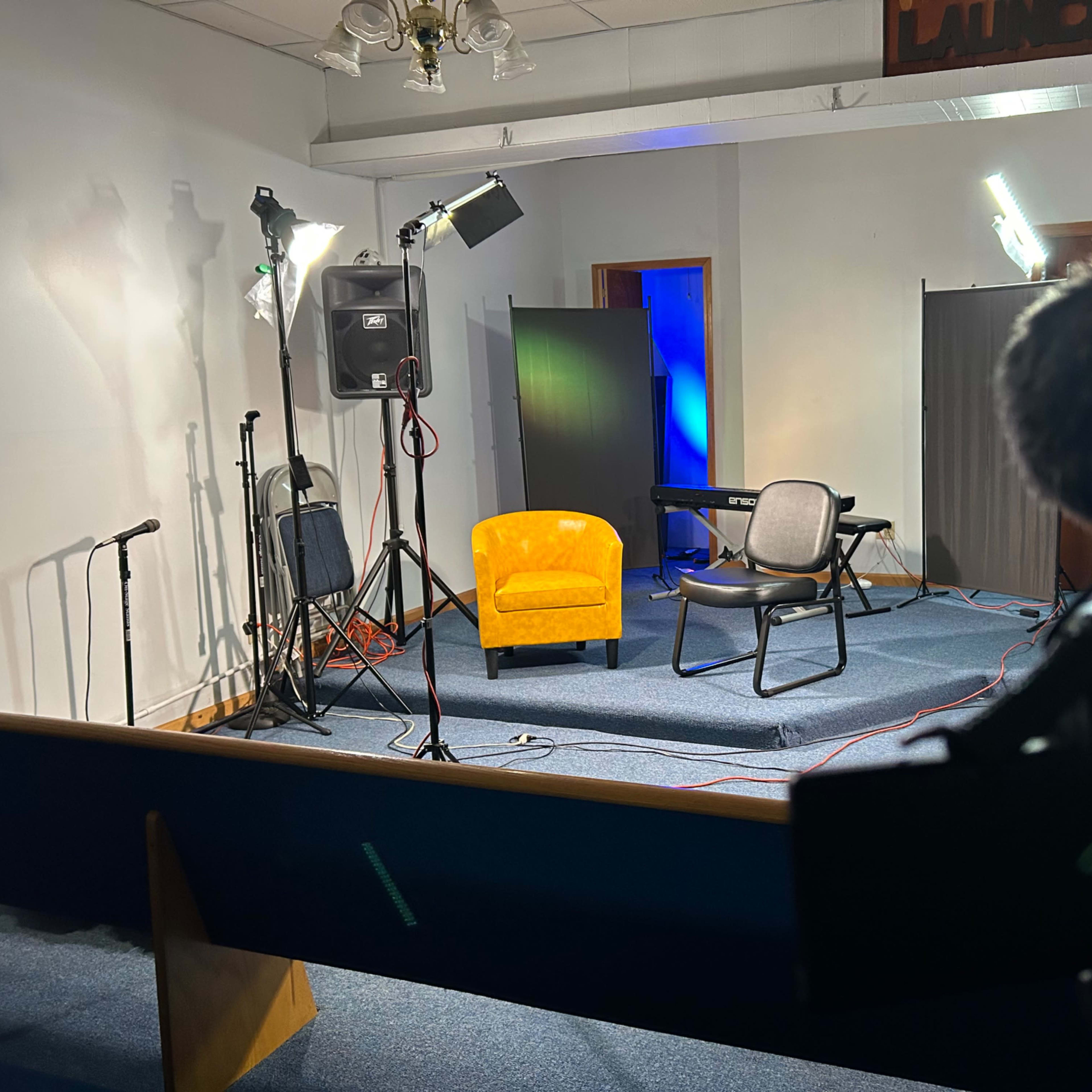 A staged interview setup with a yellow armchair, a black chair, microphones, lighting equipment, and a keyboard in a room with a blue backdrop.
