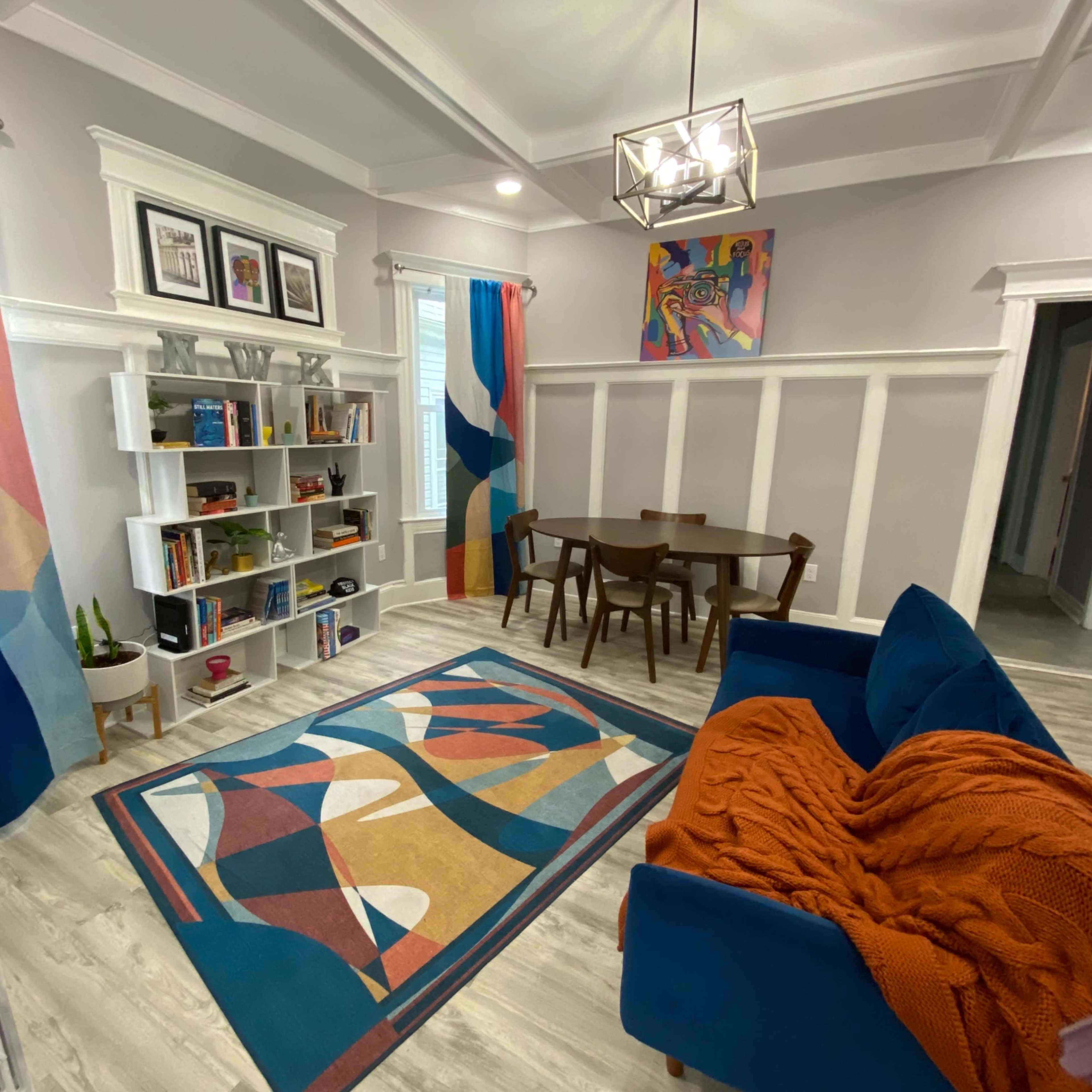 A modern living space featuring a blue couch, a patterned rug, a wooden dining table with chairs, and colorful curtains.