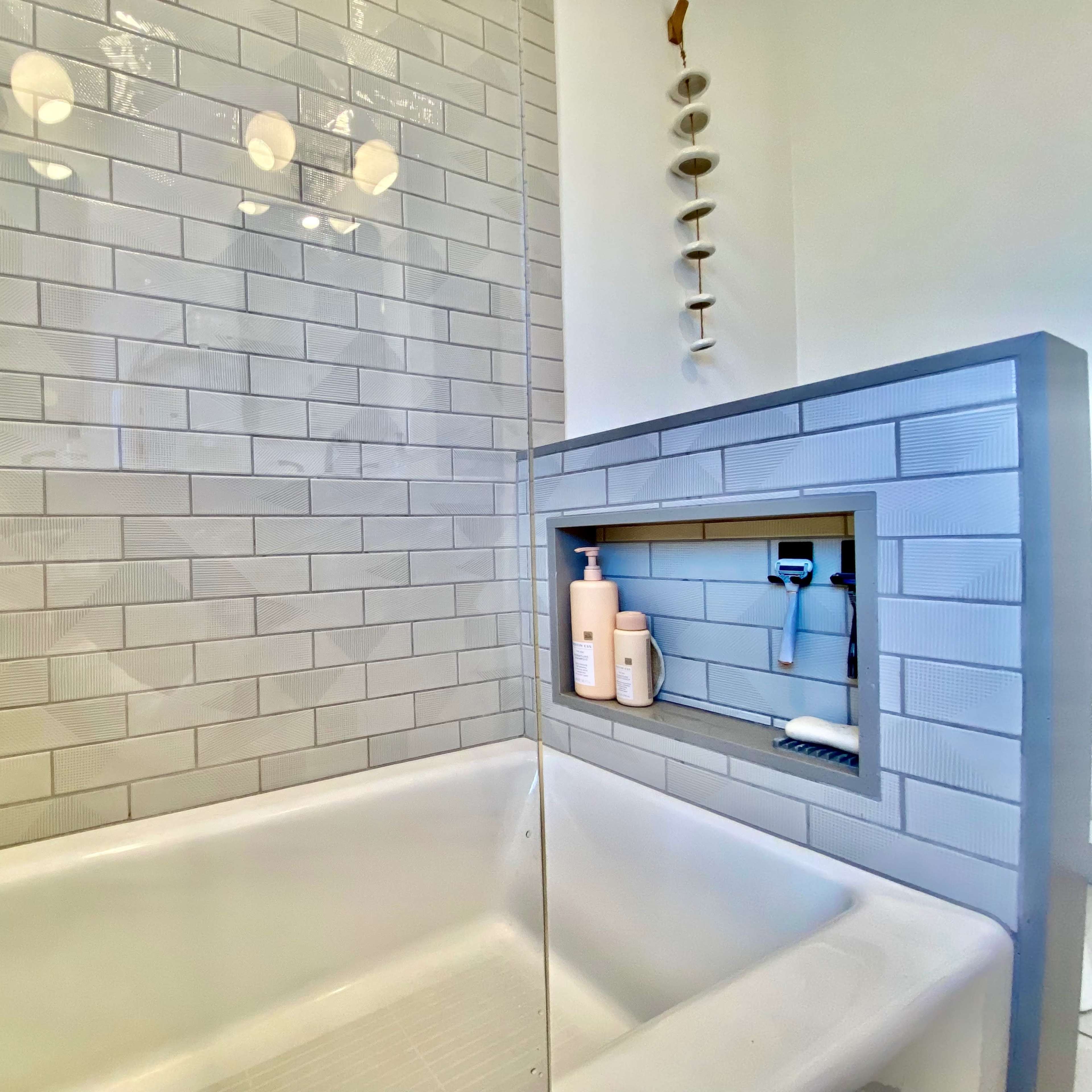 A modern bathtub enclosed by light gray tiled walls with a niche for toiletries.