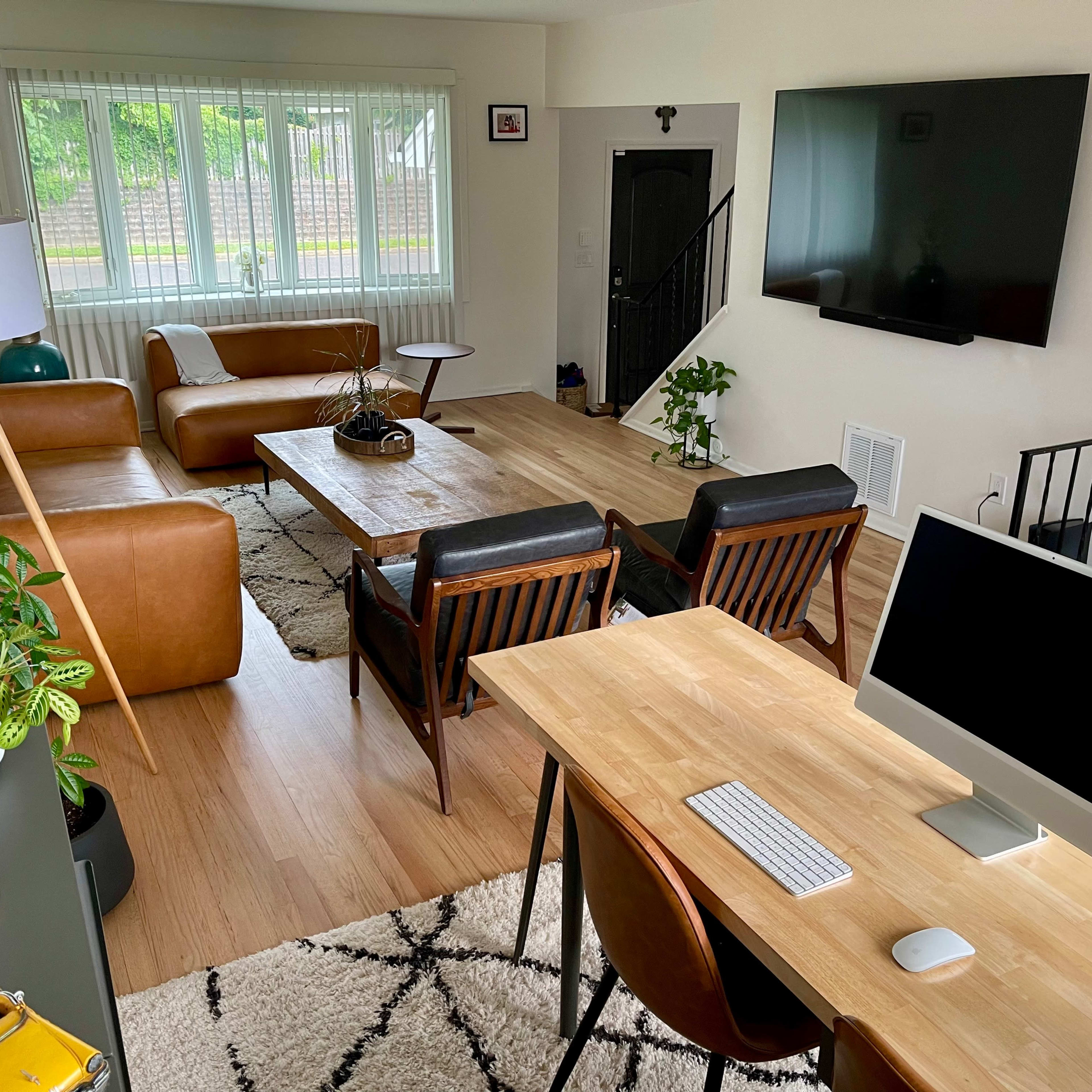 A modern living room with brown leather furniture, a wooden coffee table, a TV mounted on the wall, and a workspace featuring a computer desk.