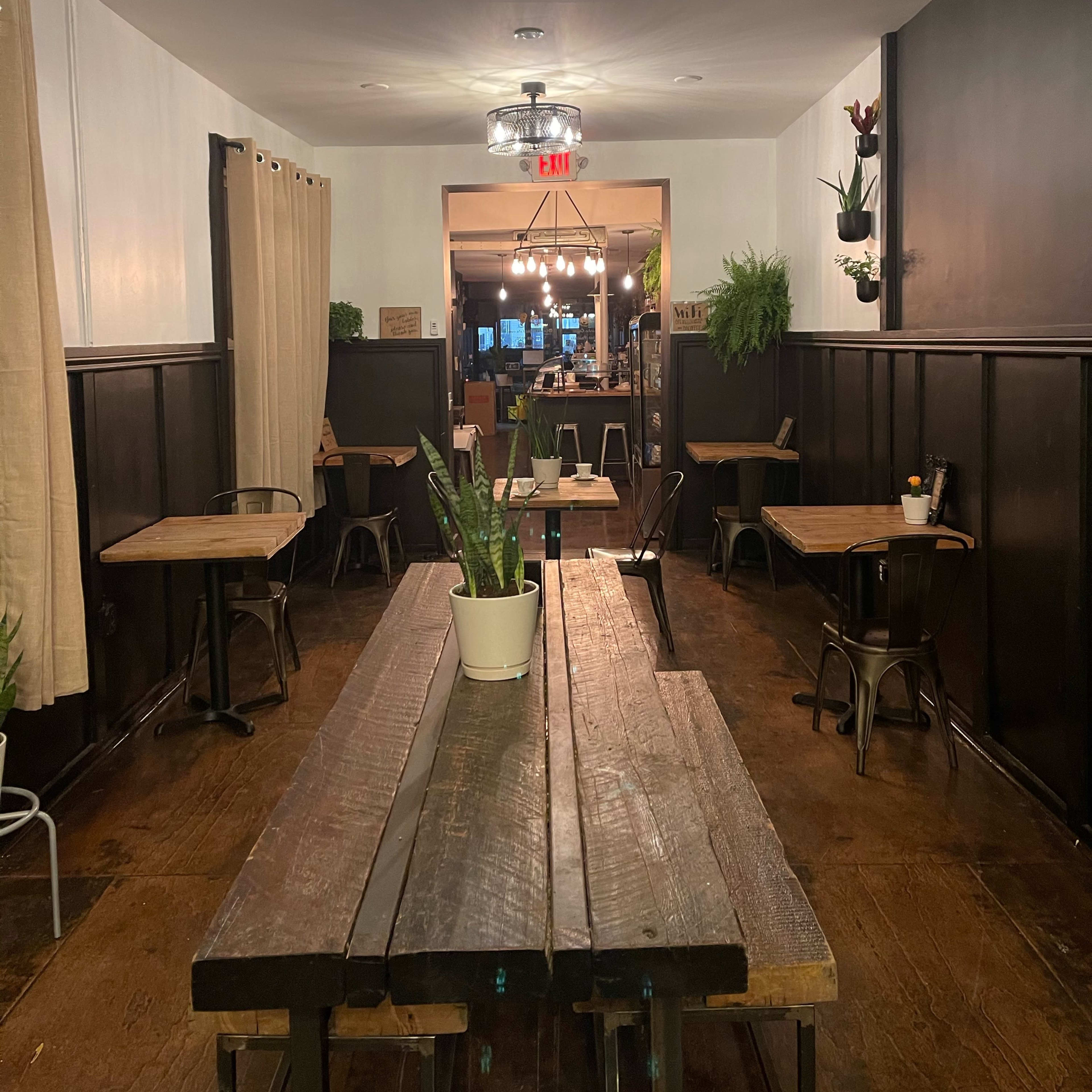 A narrow, dimly-lit restaurant hallway with wooden tables, plants, and a visible entrance to the kitchen area at the end.