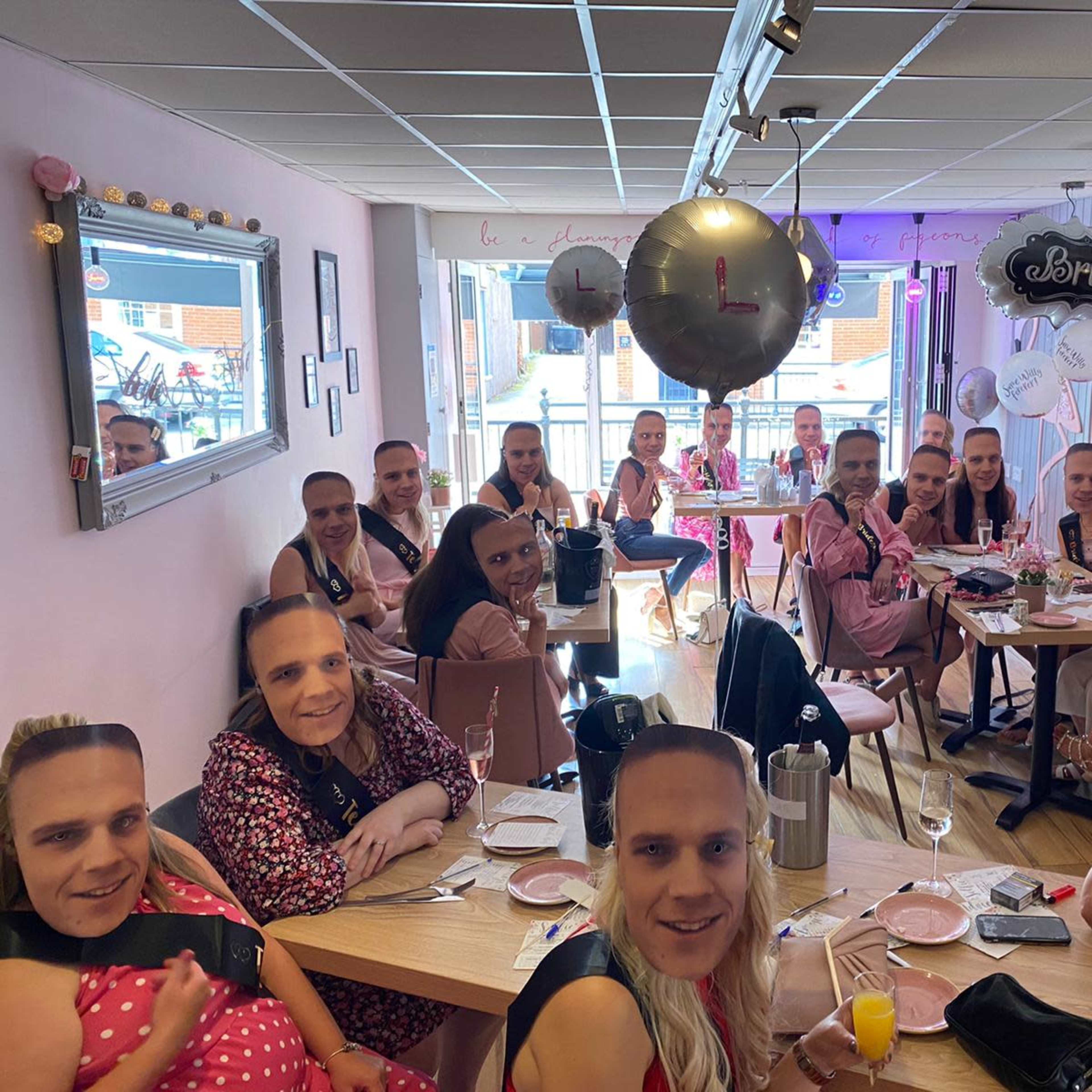 A group of people wearing masks with a man's face gathered around tables in a pink-themed restaurant for a celebration.