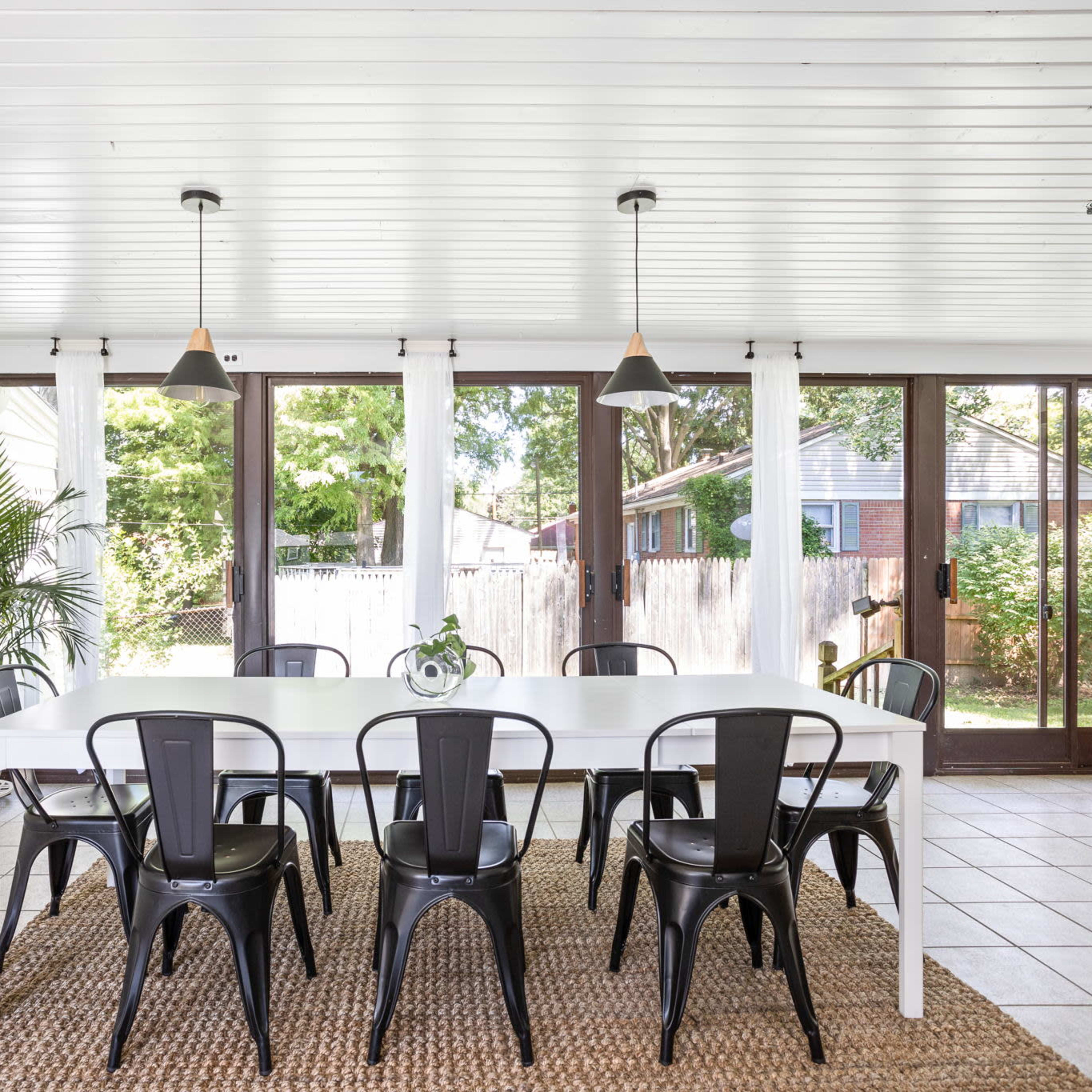 A light-filled dining area with a long white table surrounded by black chairs, large windows, and a view of a green backyard.