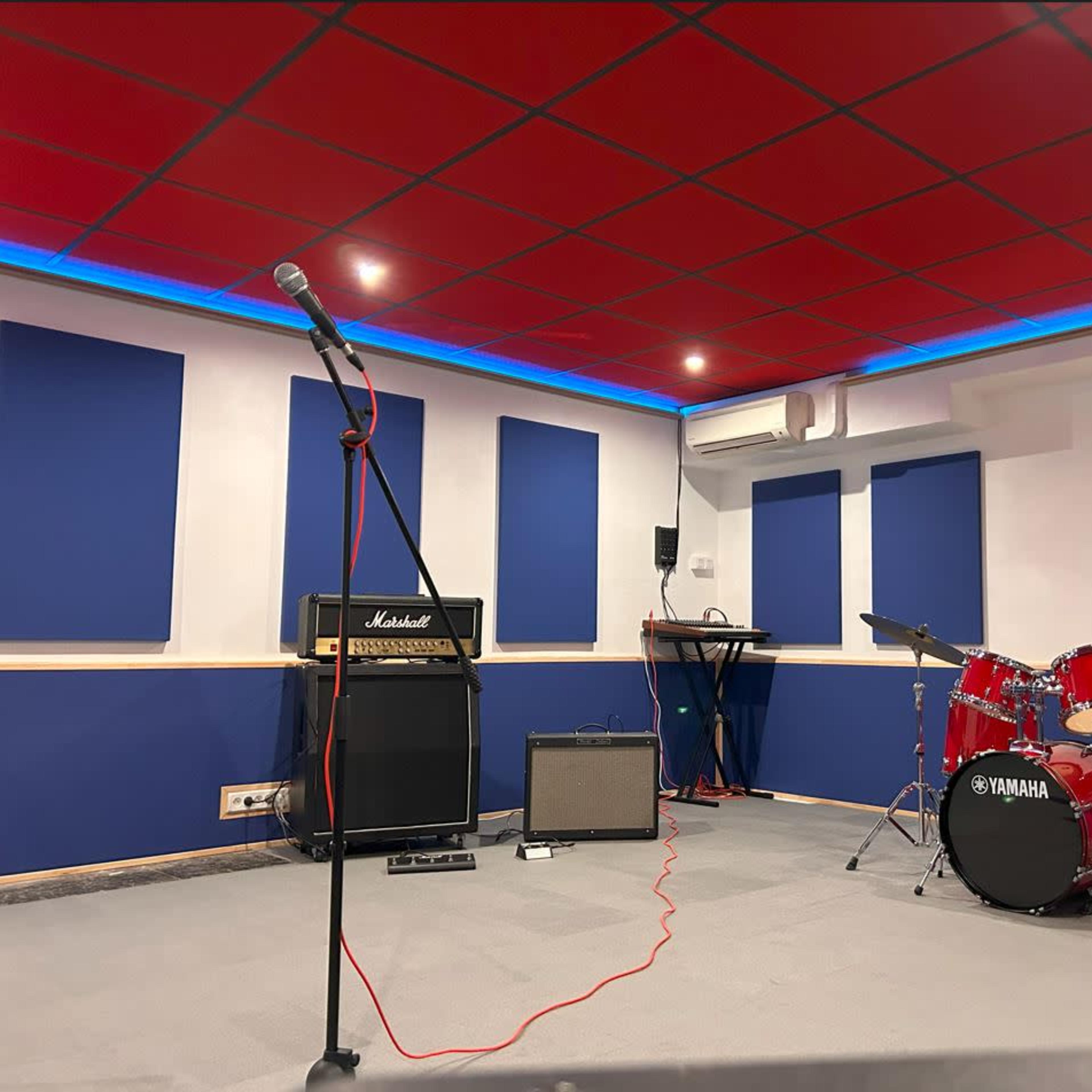A music rehearsal studio with a microphone on a stand, an amplifier, a keyboard, and a red drum set.