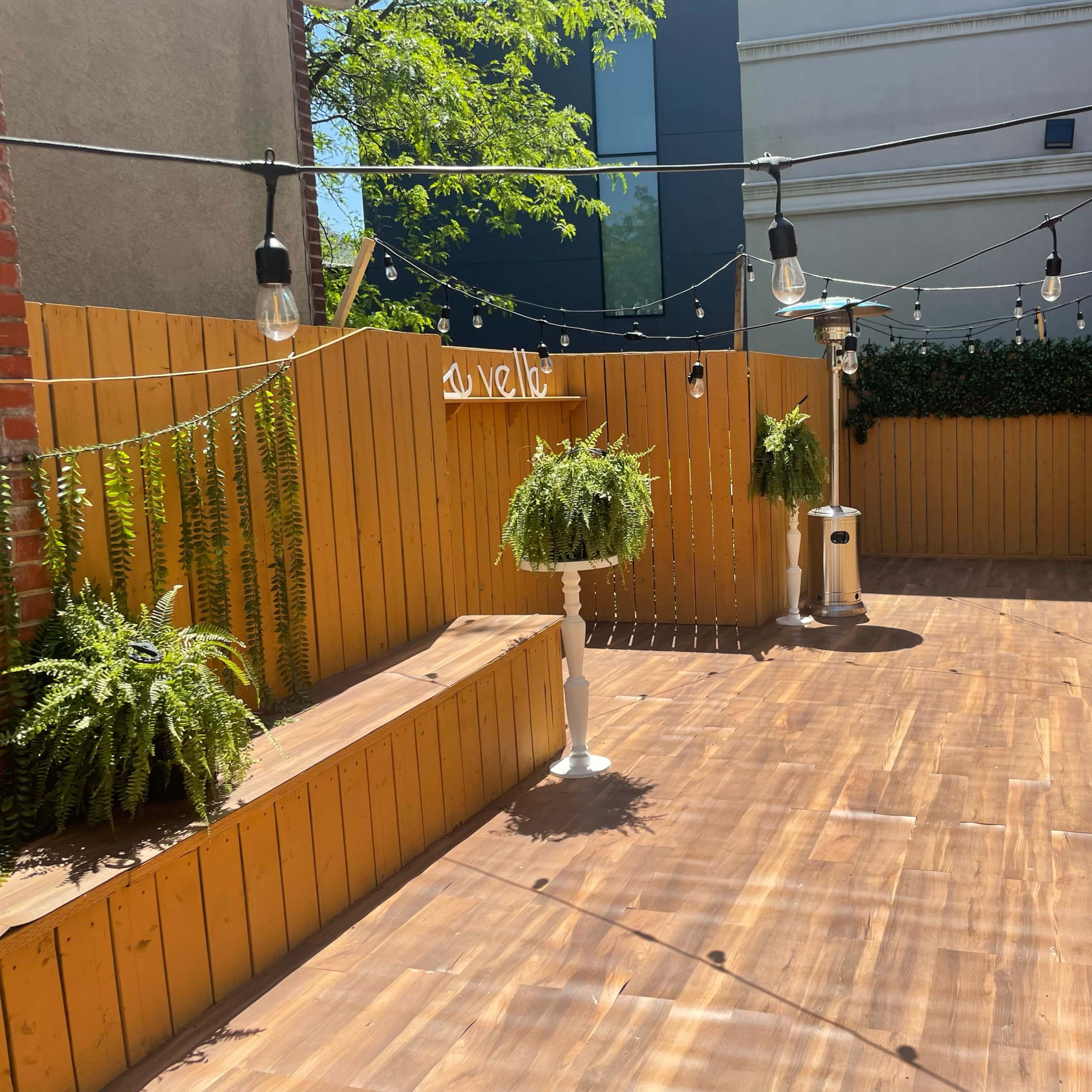A wooden outdoor space with hanging lights, potted ferns, and patio heaters surrounded by wooden fencing.
