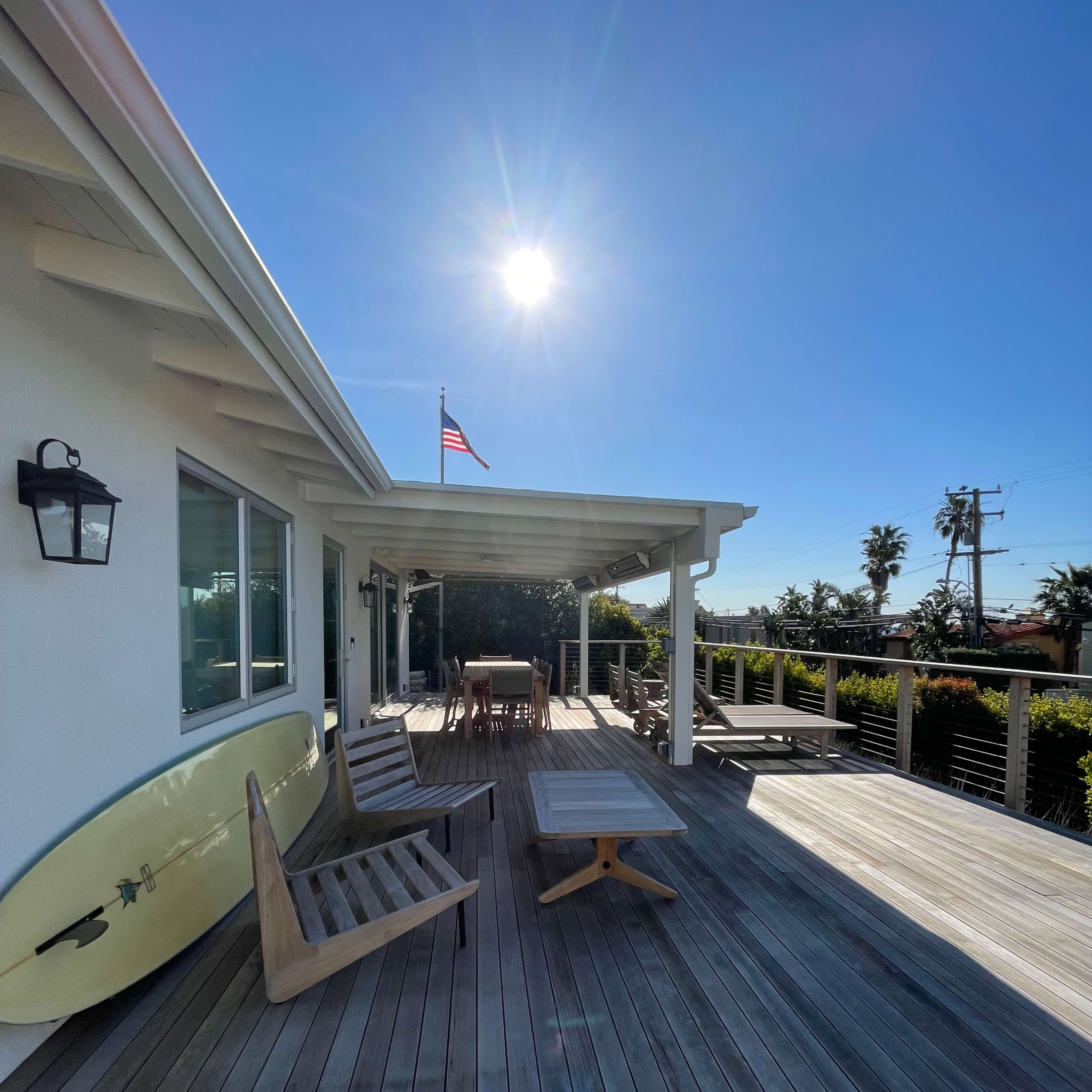 A sunlit wooden deck with a surfboard leaning against the house, outdoor furniture, and an American flag waving in the background.