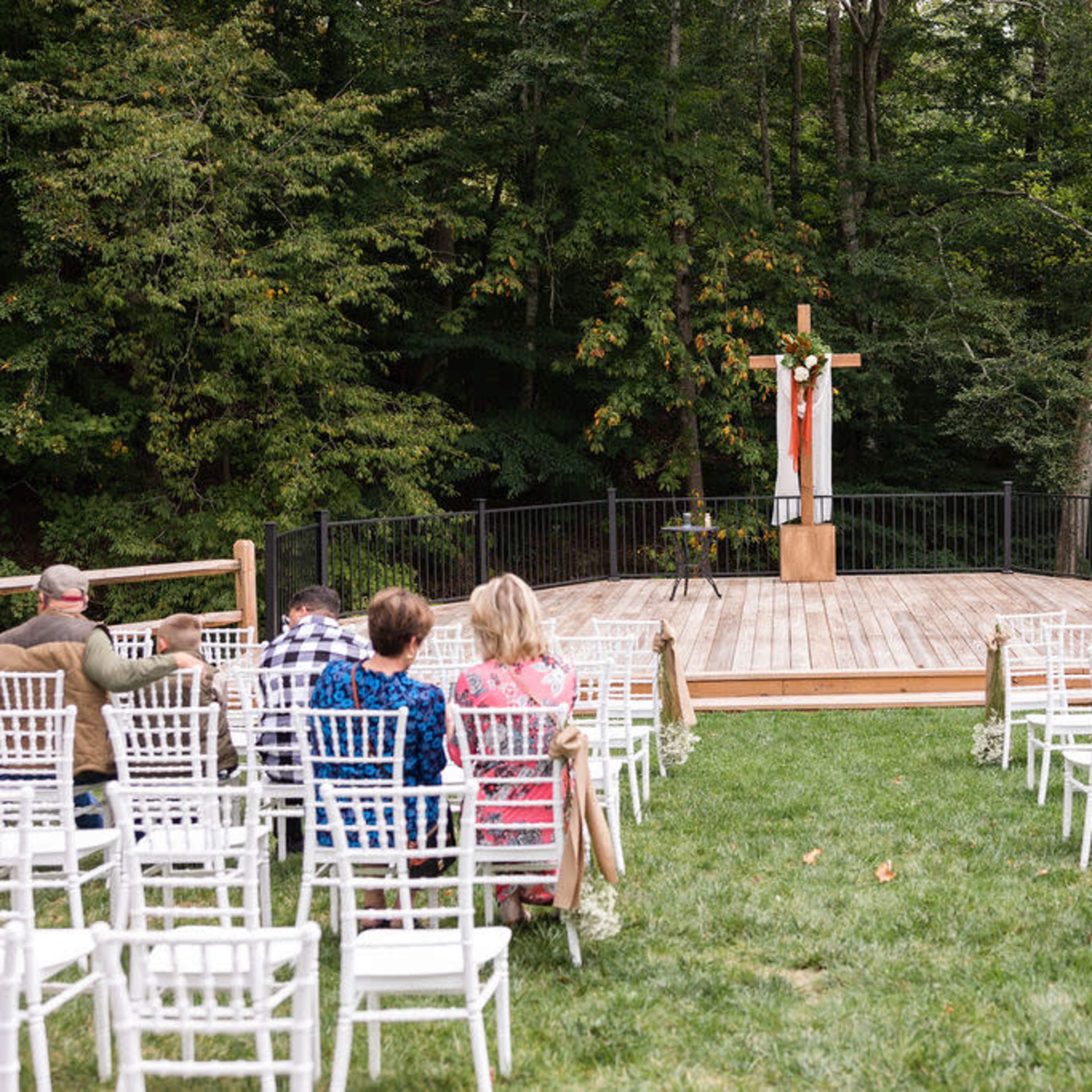 A wedding ceremony stage with a wooden platform, a cross adorned with decorations, and rows of white chairs facing it, surrounded by trees.