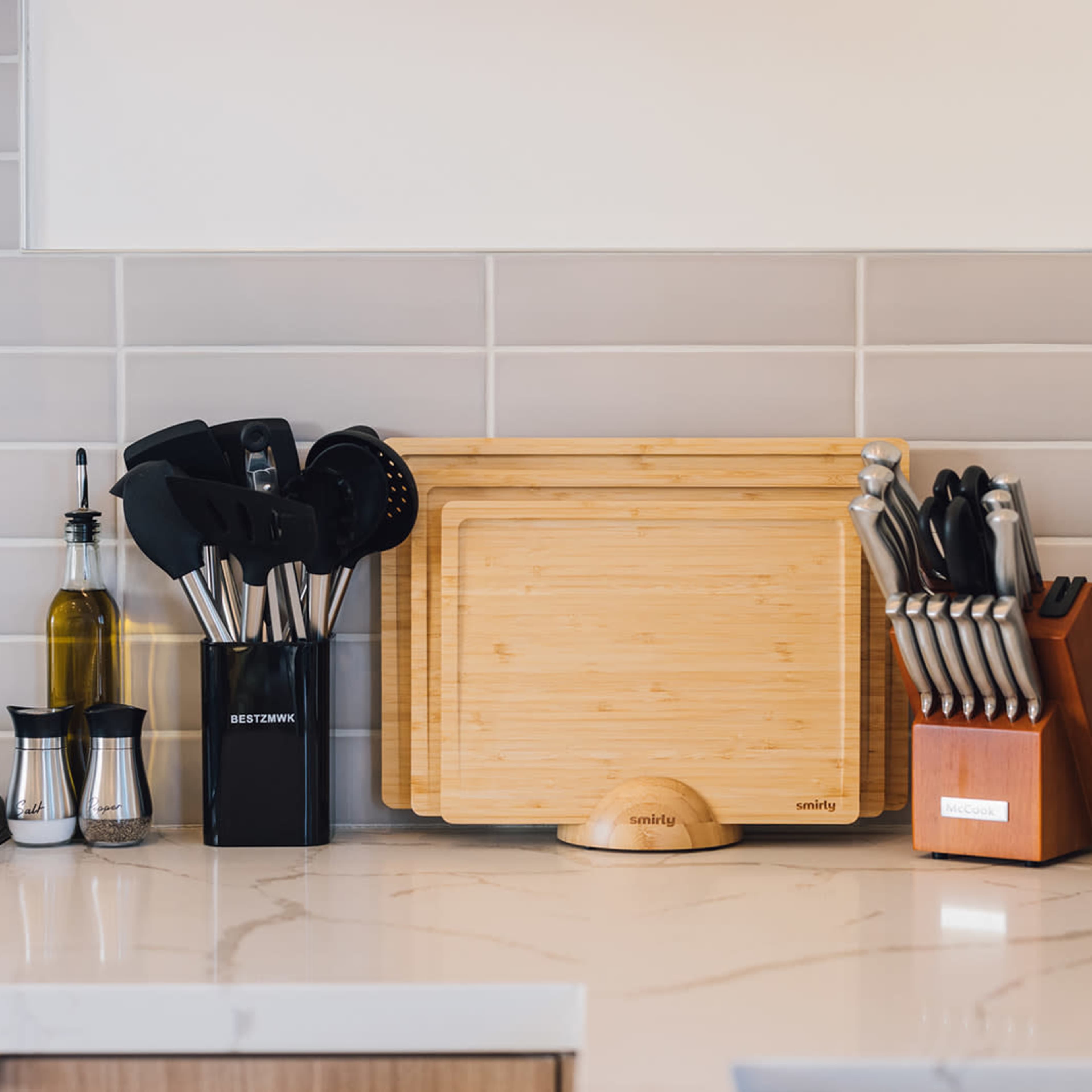 A modern kitchen countertop featuring a cutting board, various kitchen utensils in holders, a bottle of olive oil, a set of salt and pepper shakers, and a toaster.