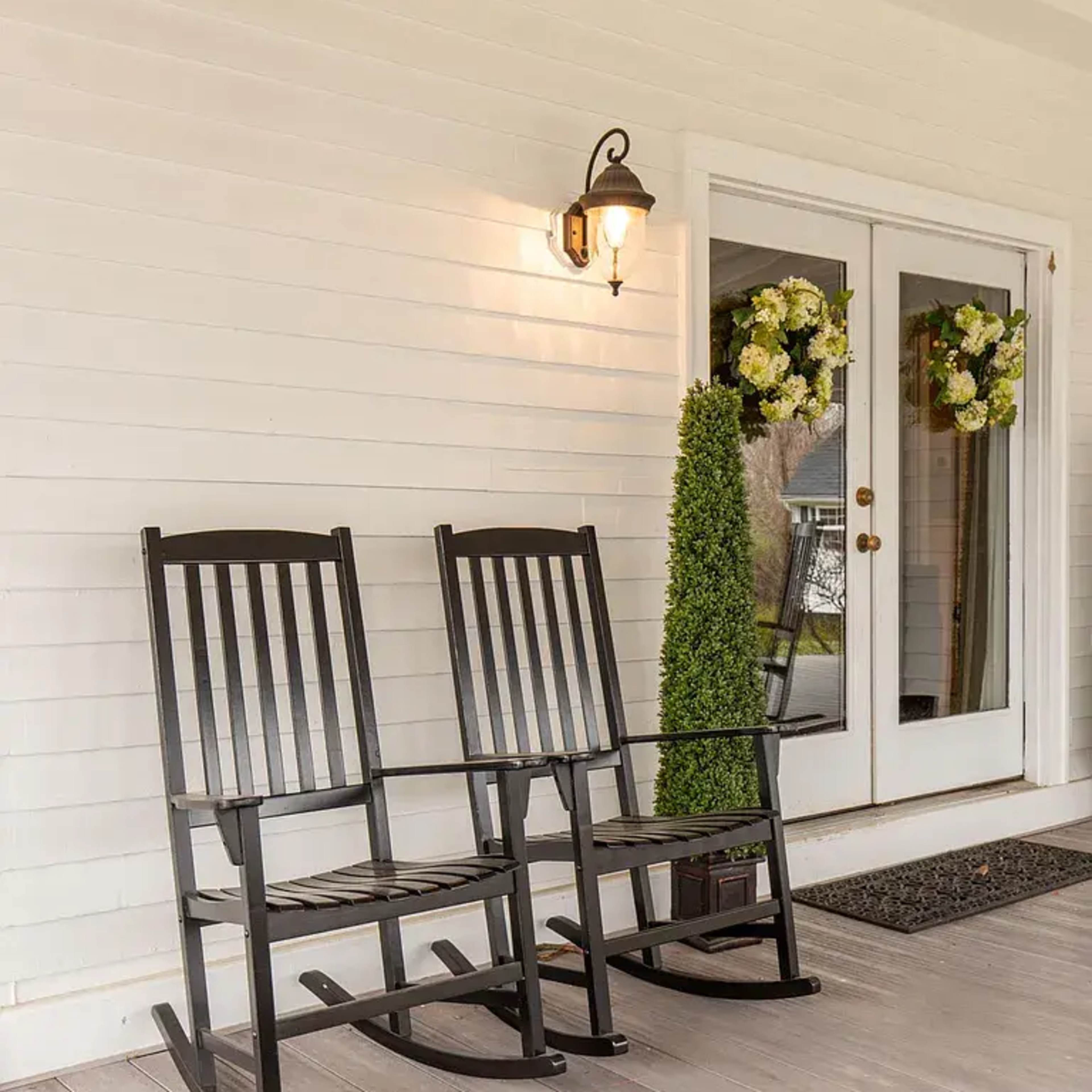 Two black rocking chairs sit on a wooden porch beside a tall green plant, with a light fixture and floral wreaths adorning the entrance.