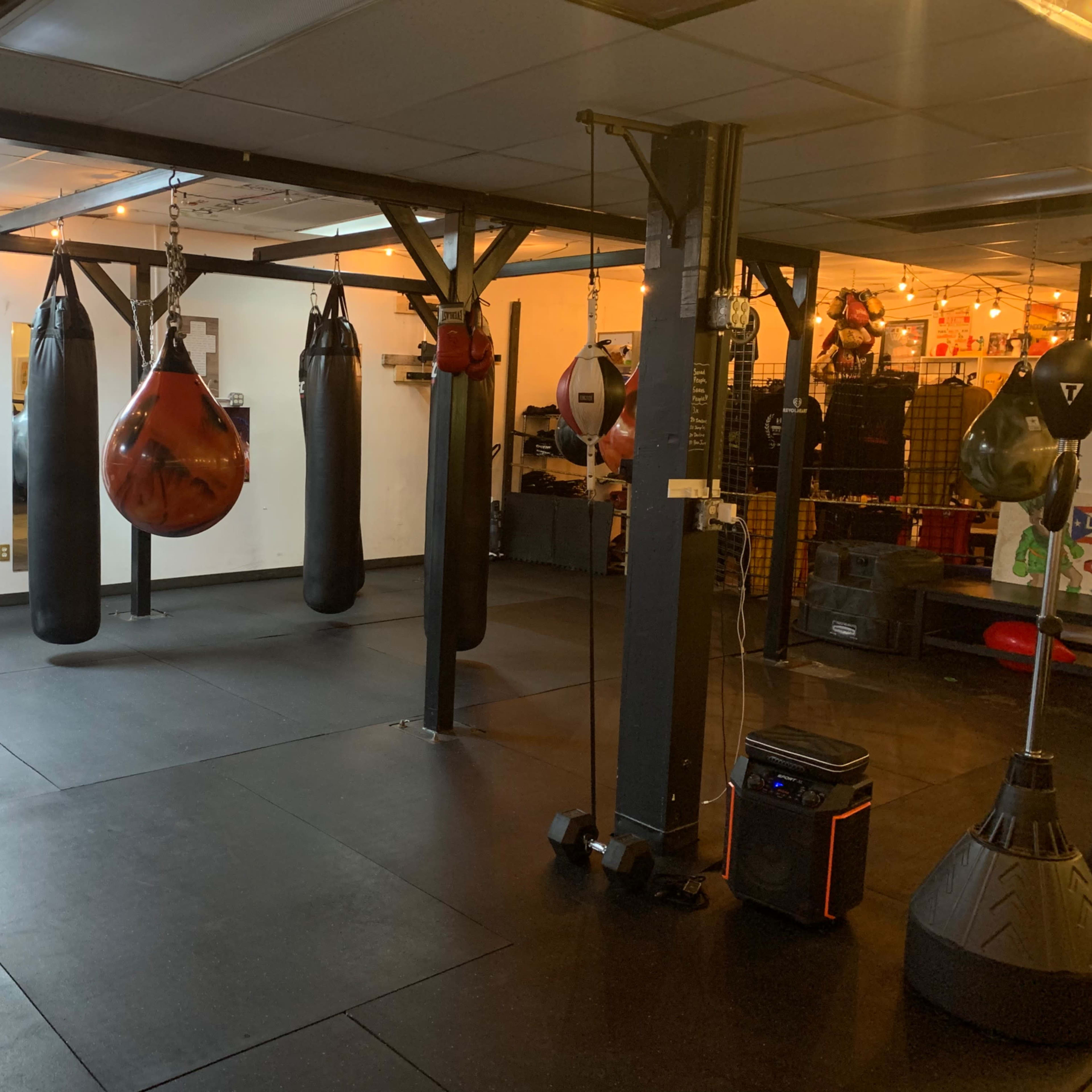 A boxing gym with several hanging punching bags, a heavy bag stand, and a black rubber flooring surface.