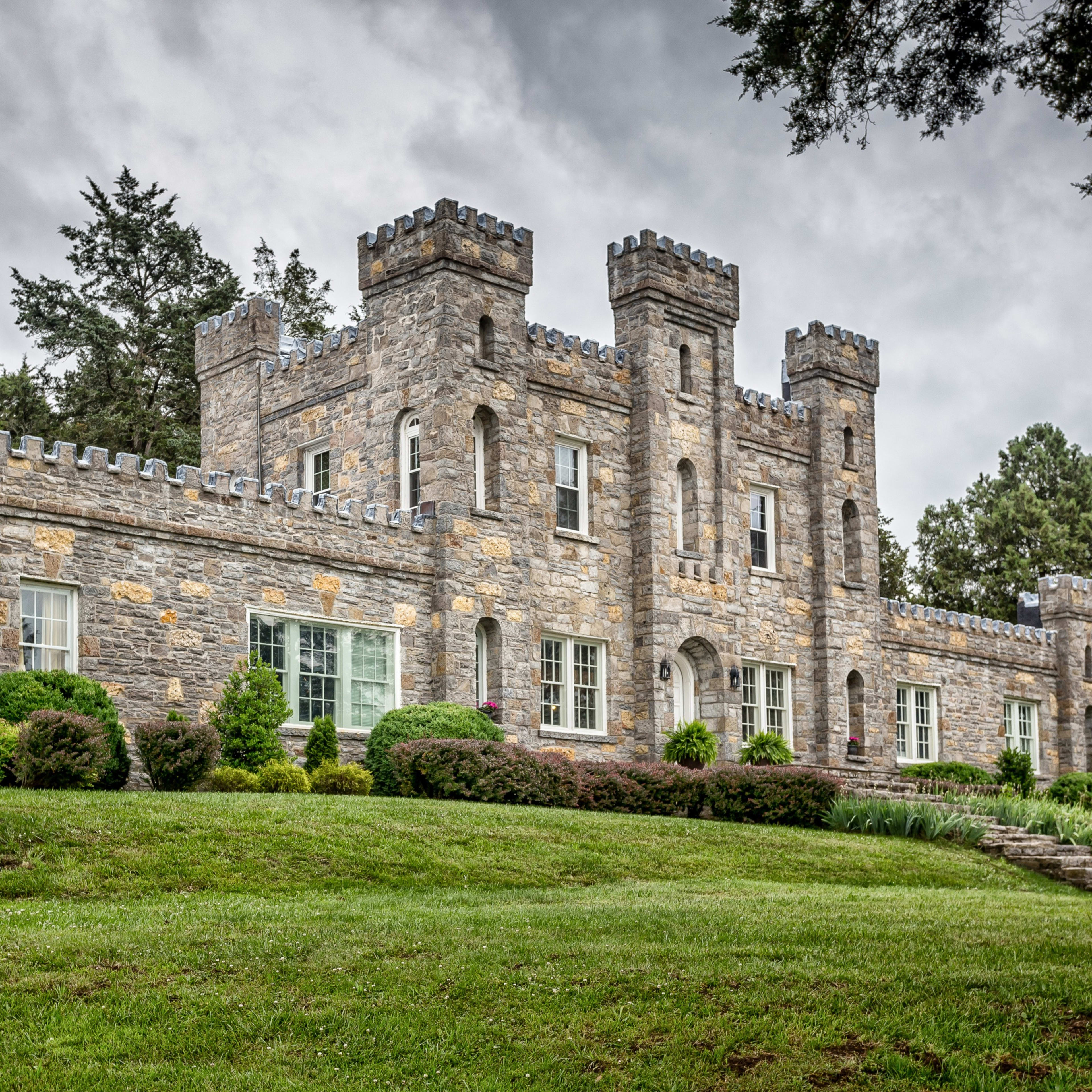 A stone castle with crenelated towers and landscaped greenery is situated on a grassy hill.