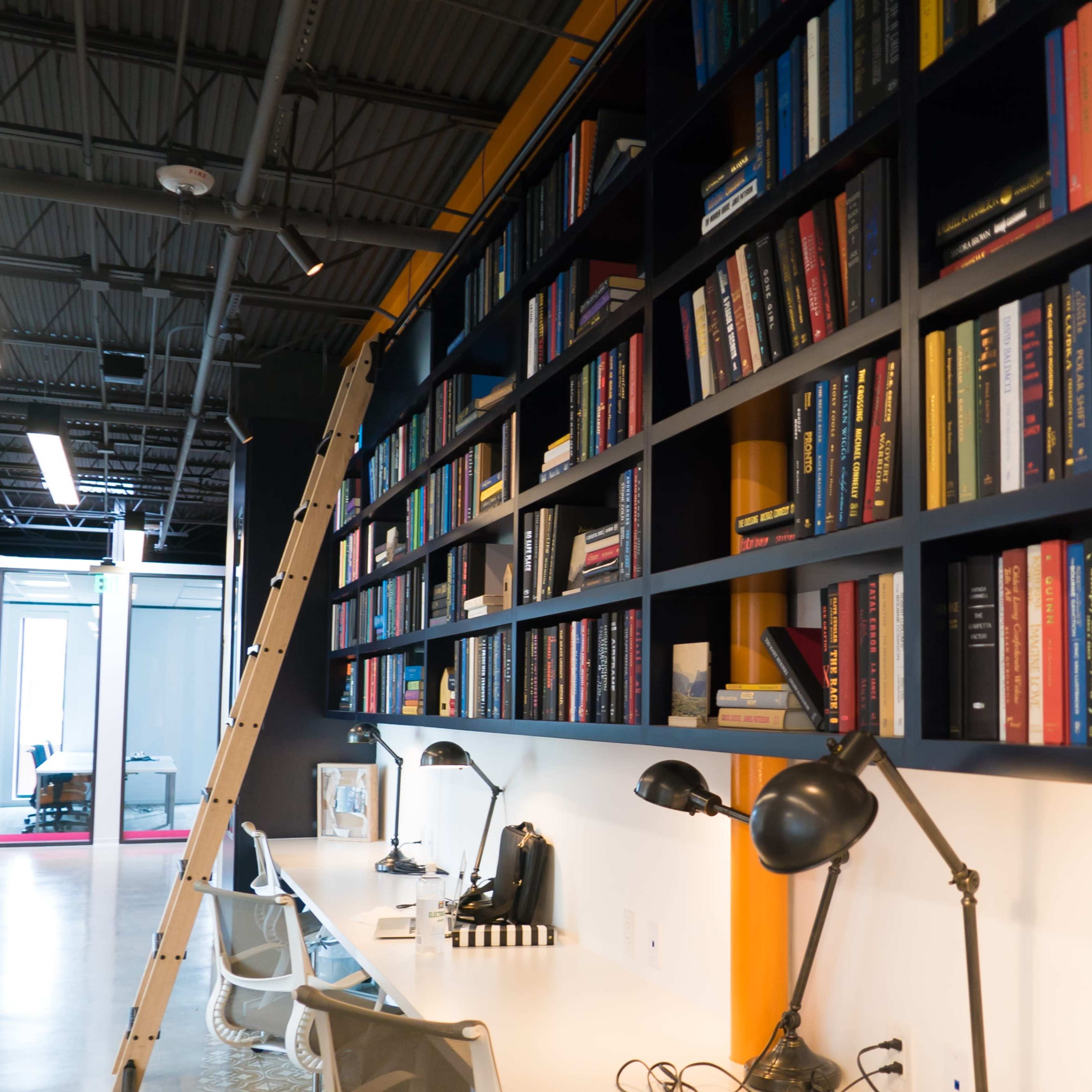 A modern office space features a long, dark shelving unit filled with books, complemented by a white desk area and a metal ladder for access.