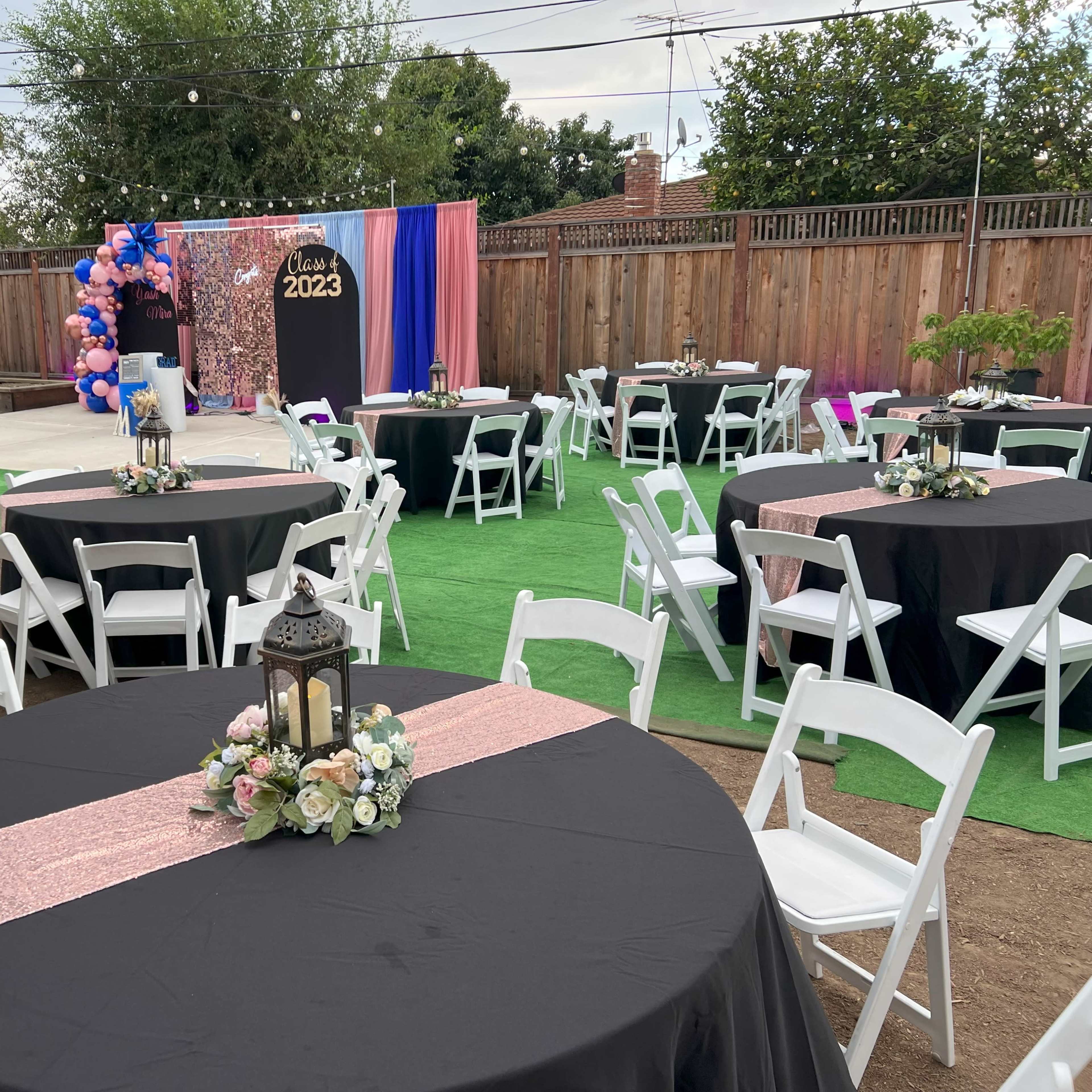 A decorated outdoor space set up for a graduation party, featuring round tables with black and pink tablecloths, lantern centerpieces, and a backdrop with balloons and a "Class of 2023" sign.