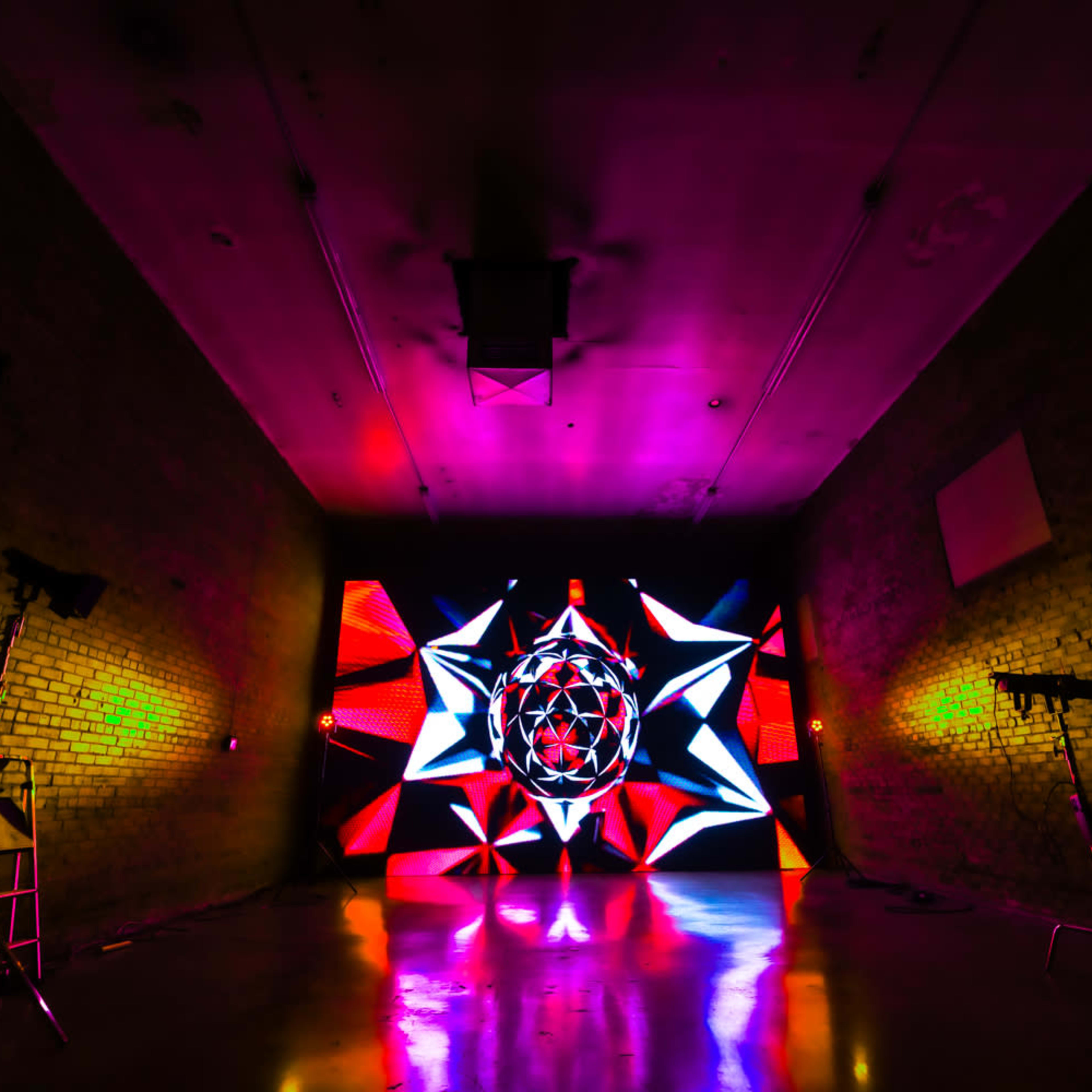 A dimly lit room with a vibrant, colorful light display featuring geometric patterns on a rear wall and stage lighting equipment set up in the corners.