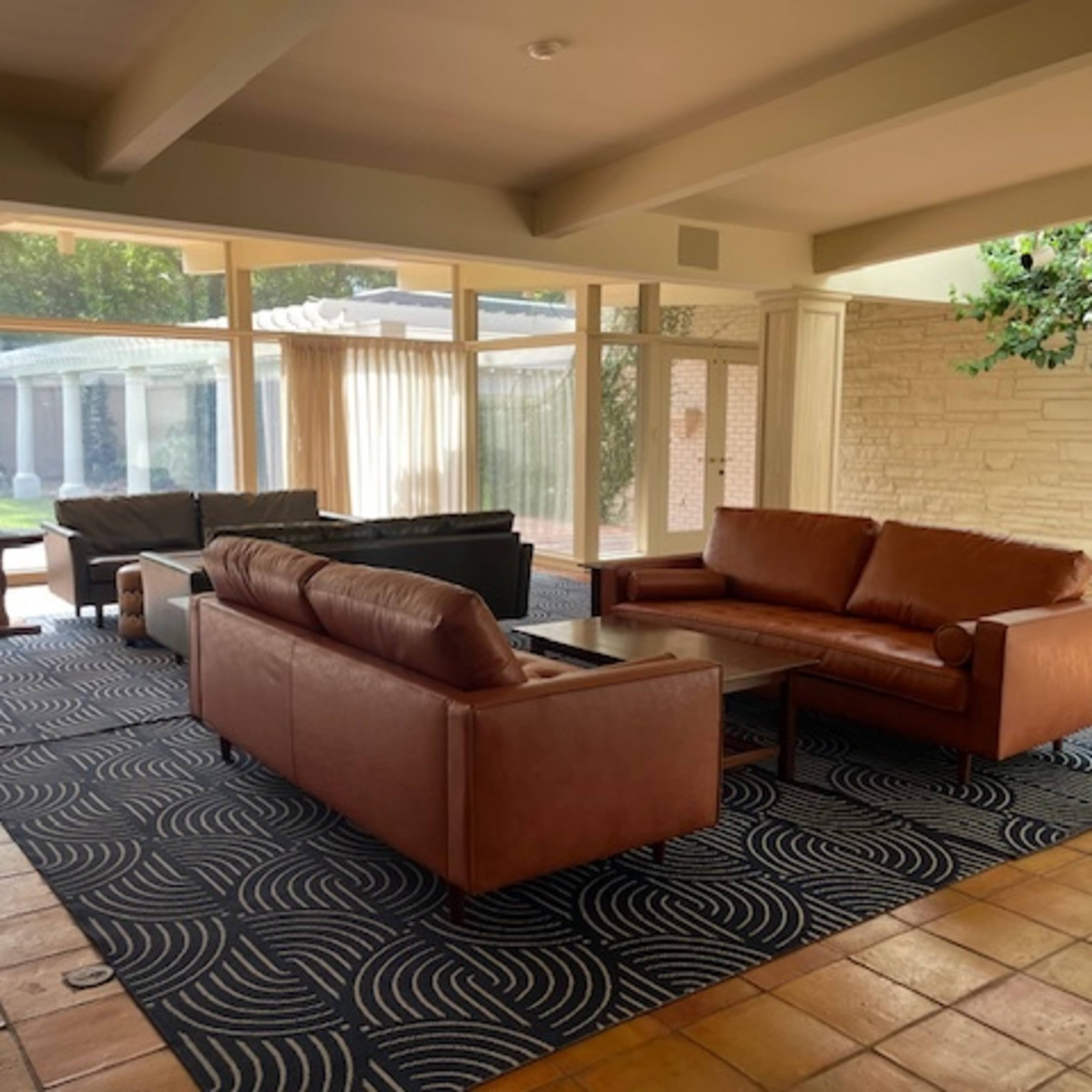 A spacious living room featuring two brown leather sofas and a coffee table on a patterned rug, with large windows allowing natural light to enter.