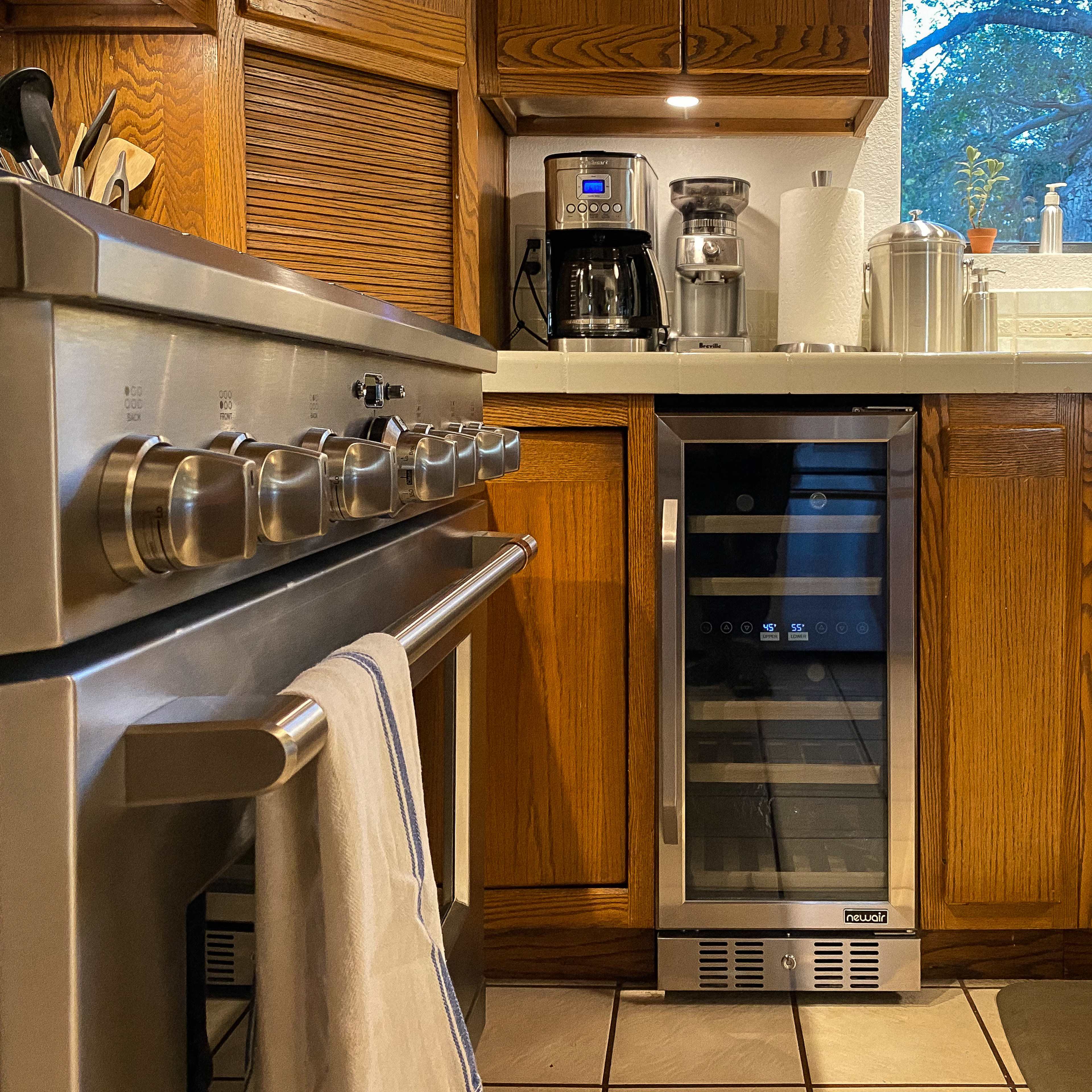 A kitchen scene features a stainless steel stove, a coffee maker, and a wine cooler set among wooden cabinets and a countertop.